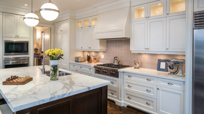 Modern kitchen with white cabinetry, marble island with sink, stainless steel appliances, vase of yellow flowers, and pendant lighting.