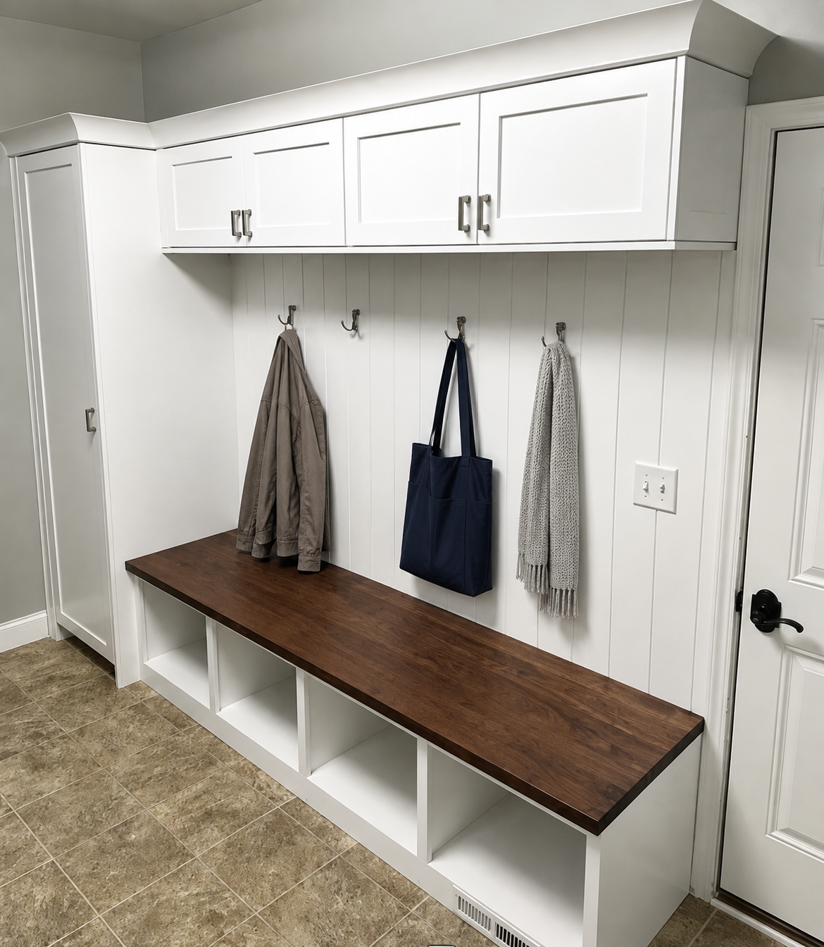 White mudroom with hooks holding jackets and scarves, a wooden bench with cubbies below, upper cabinets, and a white wall with an electrical outlet.