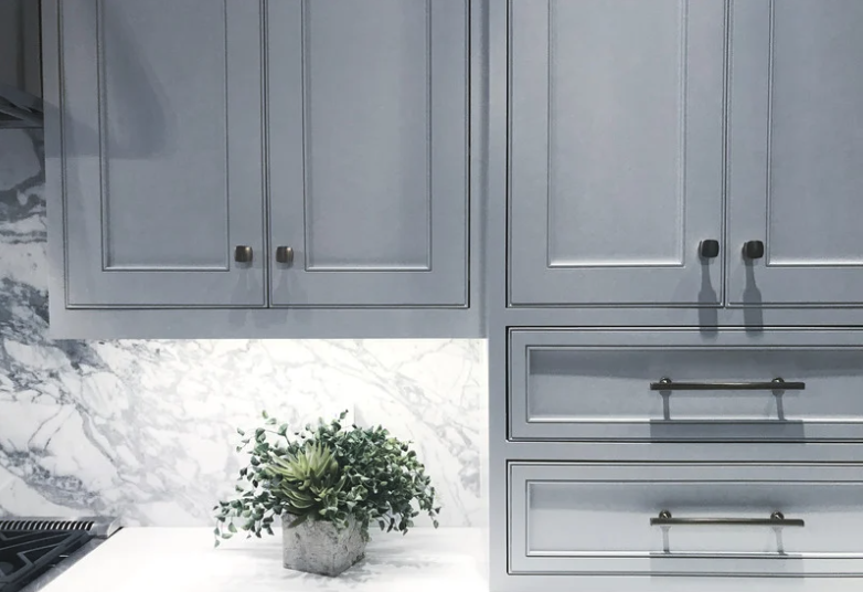 Close-up of a kitchen cabinet with light gray doors and black handles, a marble backsplash, and a potted green plant on the white marble countertop.