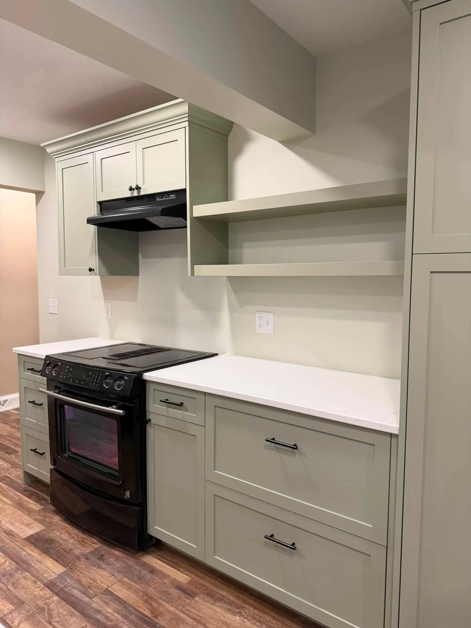 Kitchen with light green cabinets, black oven, and open wall shelves, with hardwood floor and white walls.