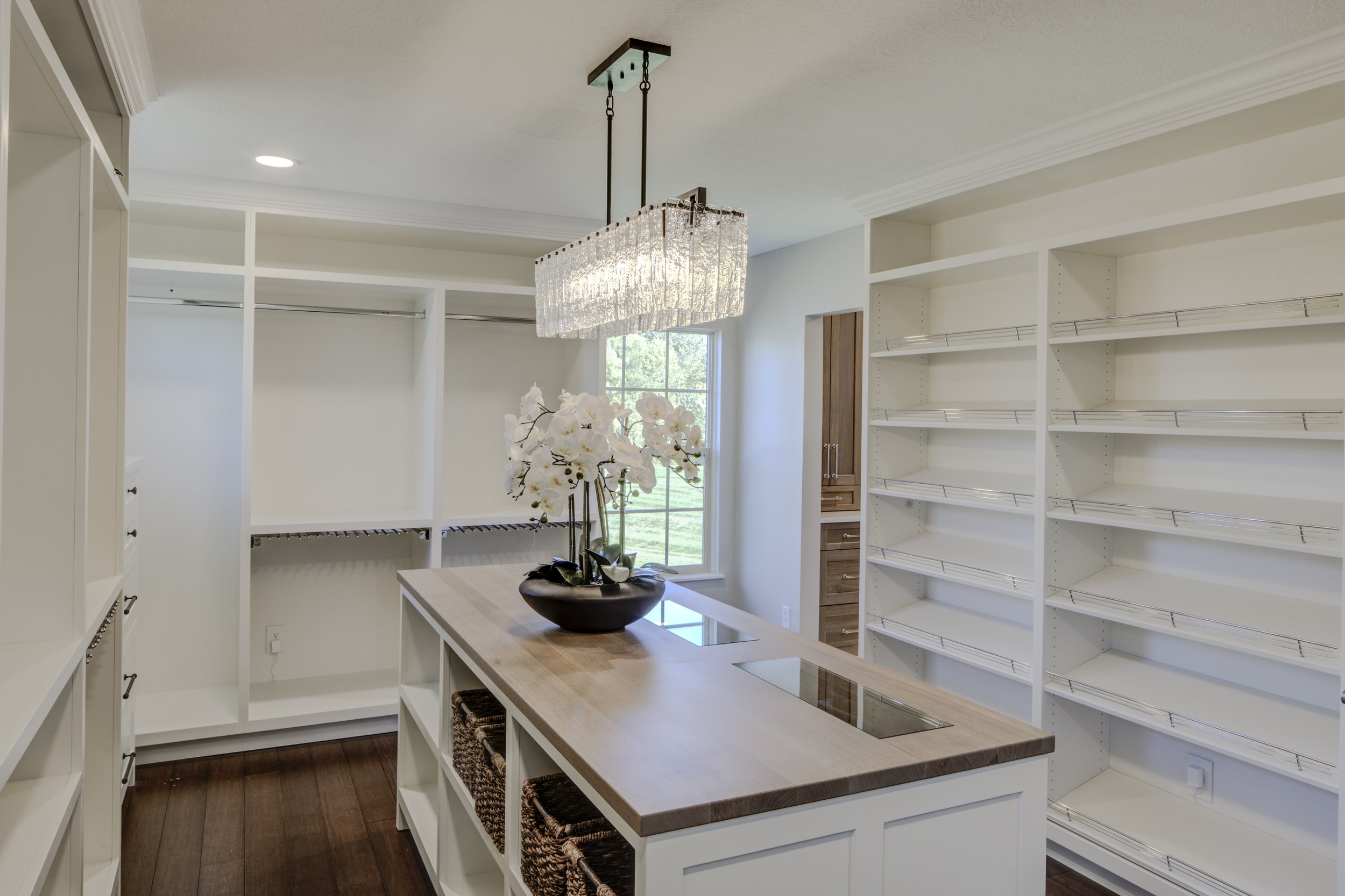 A spacious walk-in closet with white shelving units, a wooden island with baskets, a large window, a chandelier, and a potted orchid on the island.
