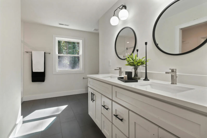 Modern bathroom with white vanity, two black-framed oval mirrors, black light fixtures, black drawer handles, and a window letting in sunlight.