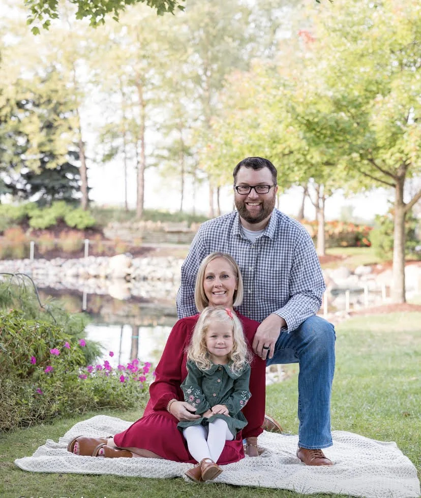Family of three outside in a park with trees, pond, and flowers. The man is kneeling with a smile, the woman is sitting on a blanket with a girl in a green dress sitting in front.