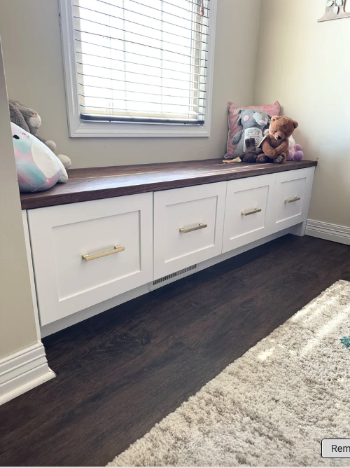 White built-in window seat with a wooden top, three drawers with gold handles underneath, plush toys including a teddy bear and a stuffed animal, a window with blinds, and a section of beige wall with a small part of a picture frame visible.