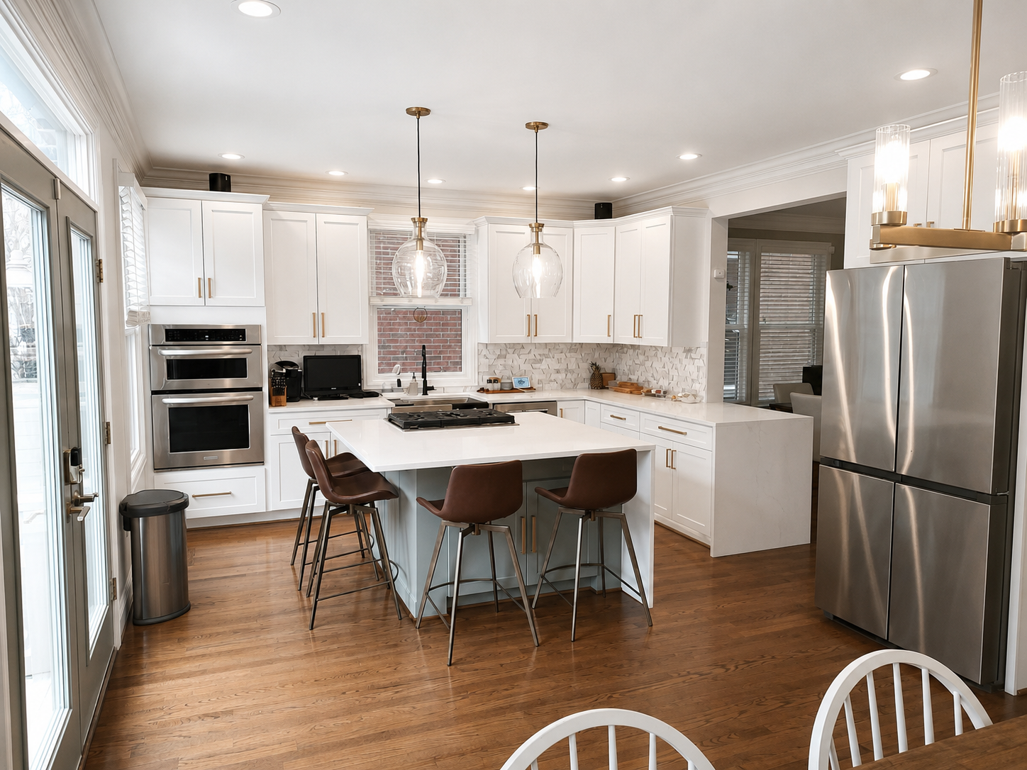 Modern kitchen with white cabinets, stainless steel refrigerator, double oven, island with brown chairs, wooden floor, pendant lights, and natural light from glass doors.