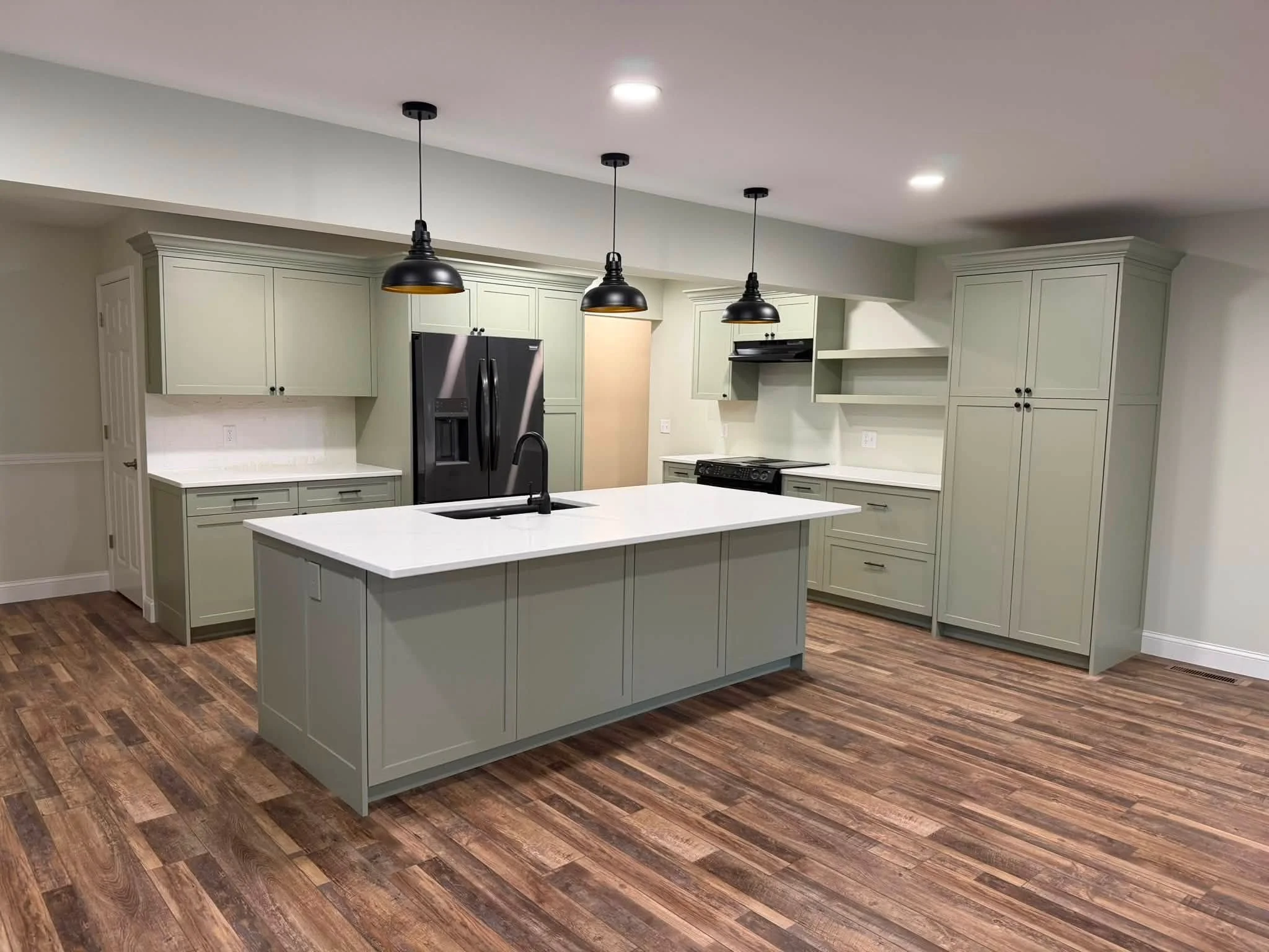 A modern kitchen with light green cabinets, black pendant lights, a white kitchen island, stainless steel fridge, black stove, and wood flooring.