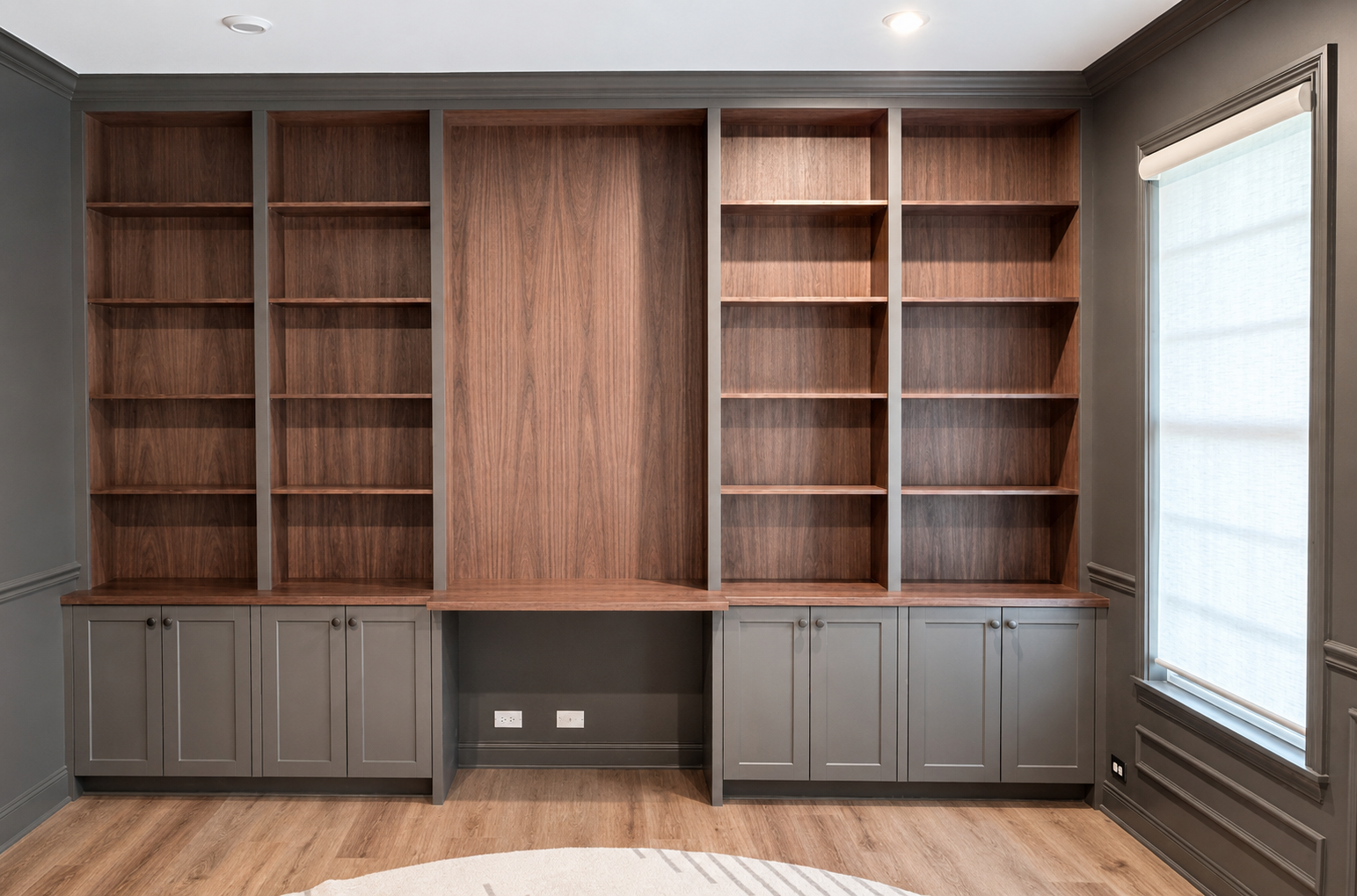 Empty built-in wooden bookshelf with gray cabinet doors underneath, located in a room with wood flooring and gray walls.