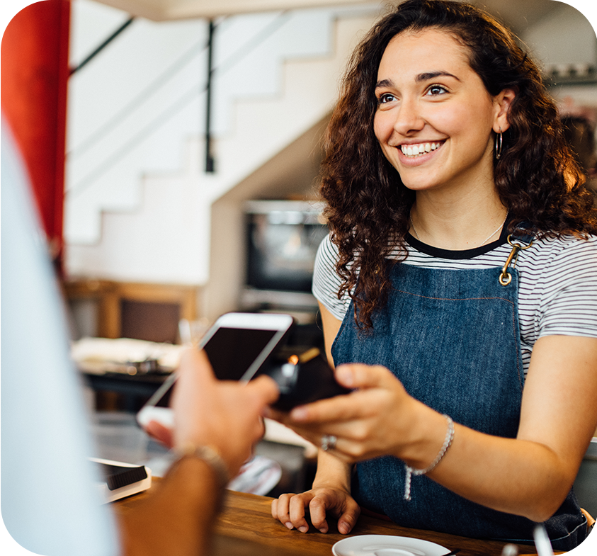 Smiling barista in denim apron interacting with customer holding smartphone at cafe counter.