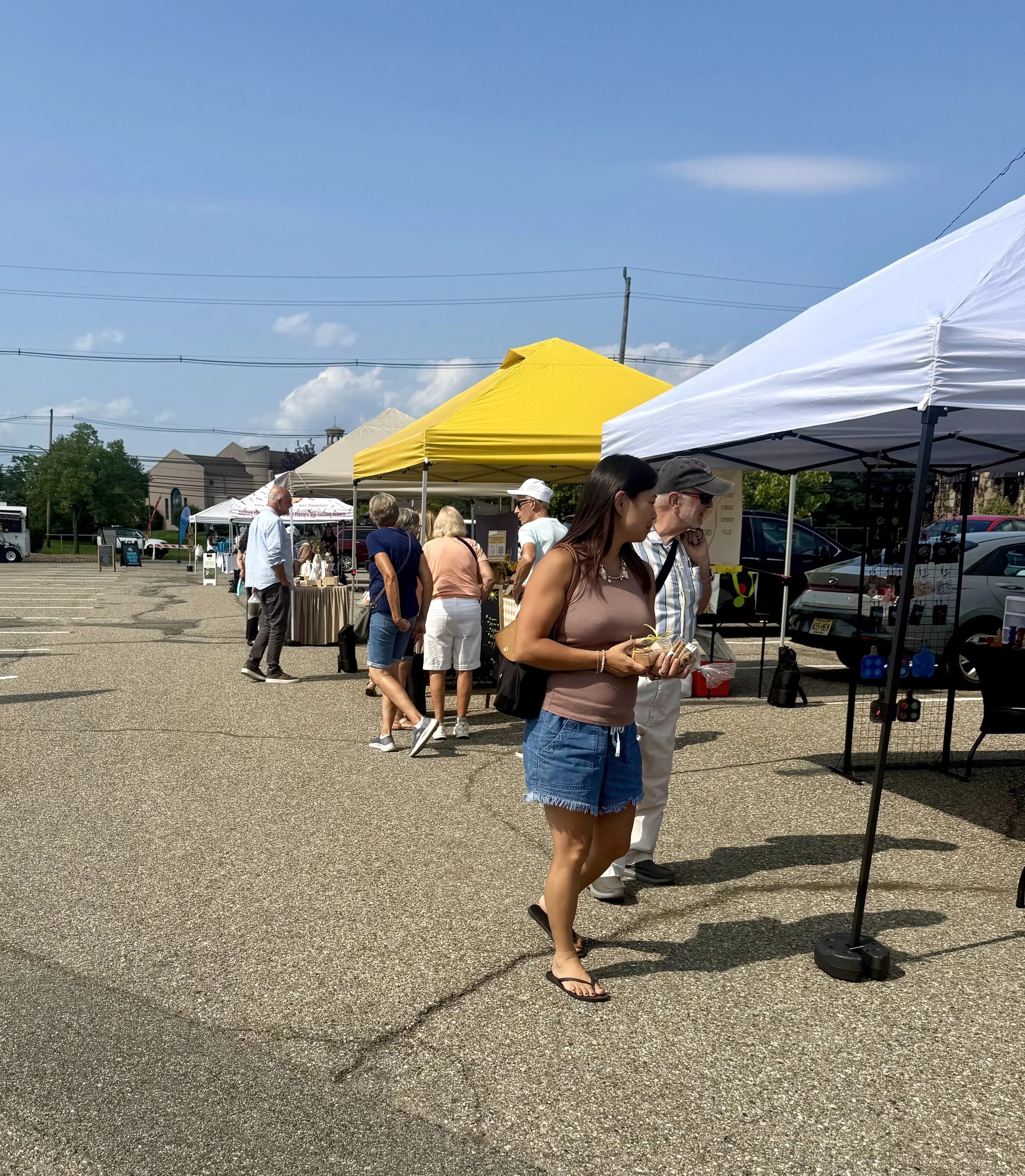 People browsing outdoor market stalls under white and yellow canopies on a sunny day with a parking lot and buildings in the background. Parsippany Farmers market by Hear Here Events 2026 season