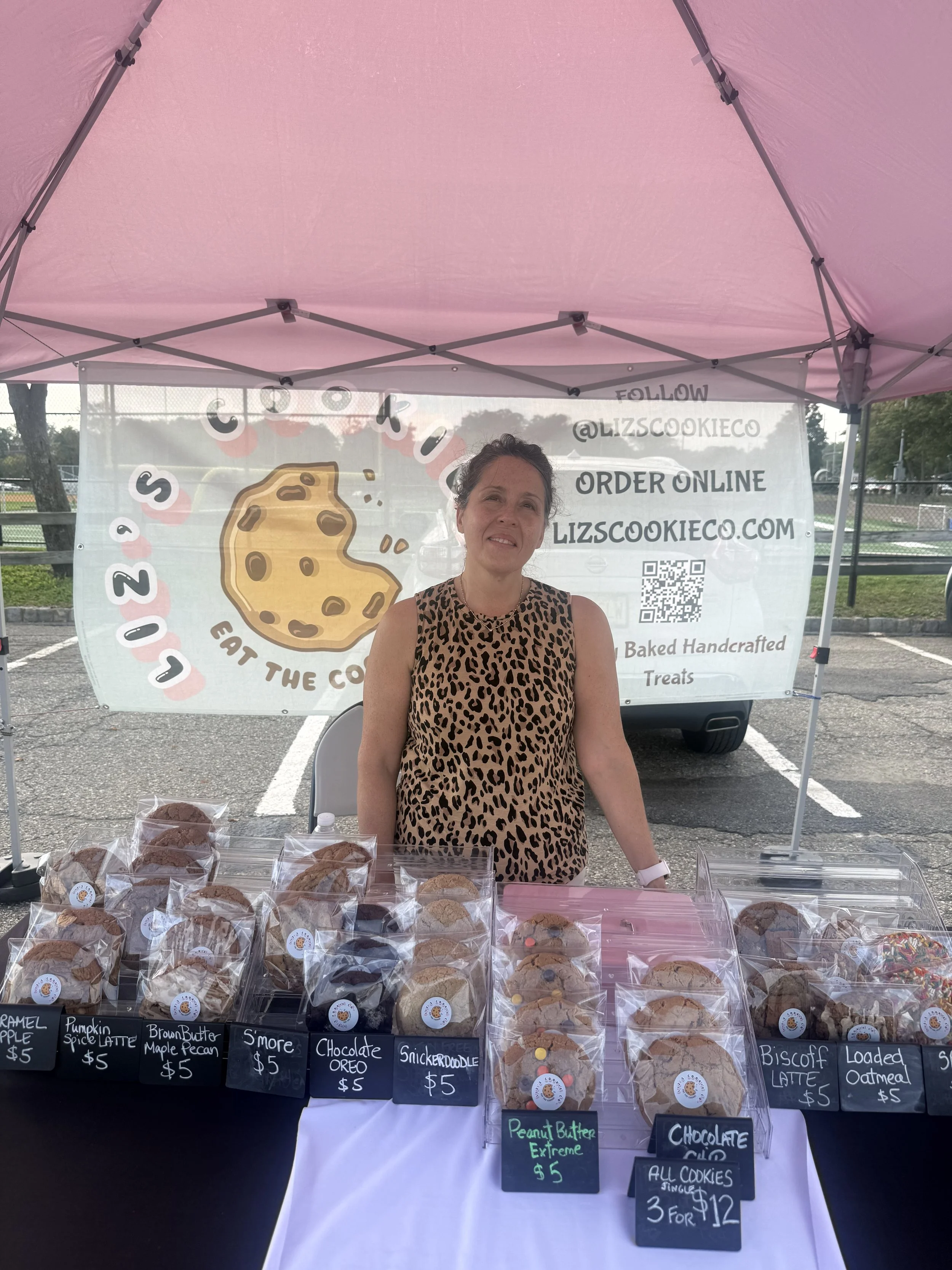 A woman standing behind a table of baked cookies at an outdoor market stall, with signs displaying various cookie flavors and prices. Behind her, a promotional banner for 'Cookie' brand cookies,Parsippany Farmers market by Hear Here Events 2026 