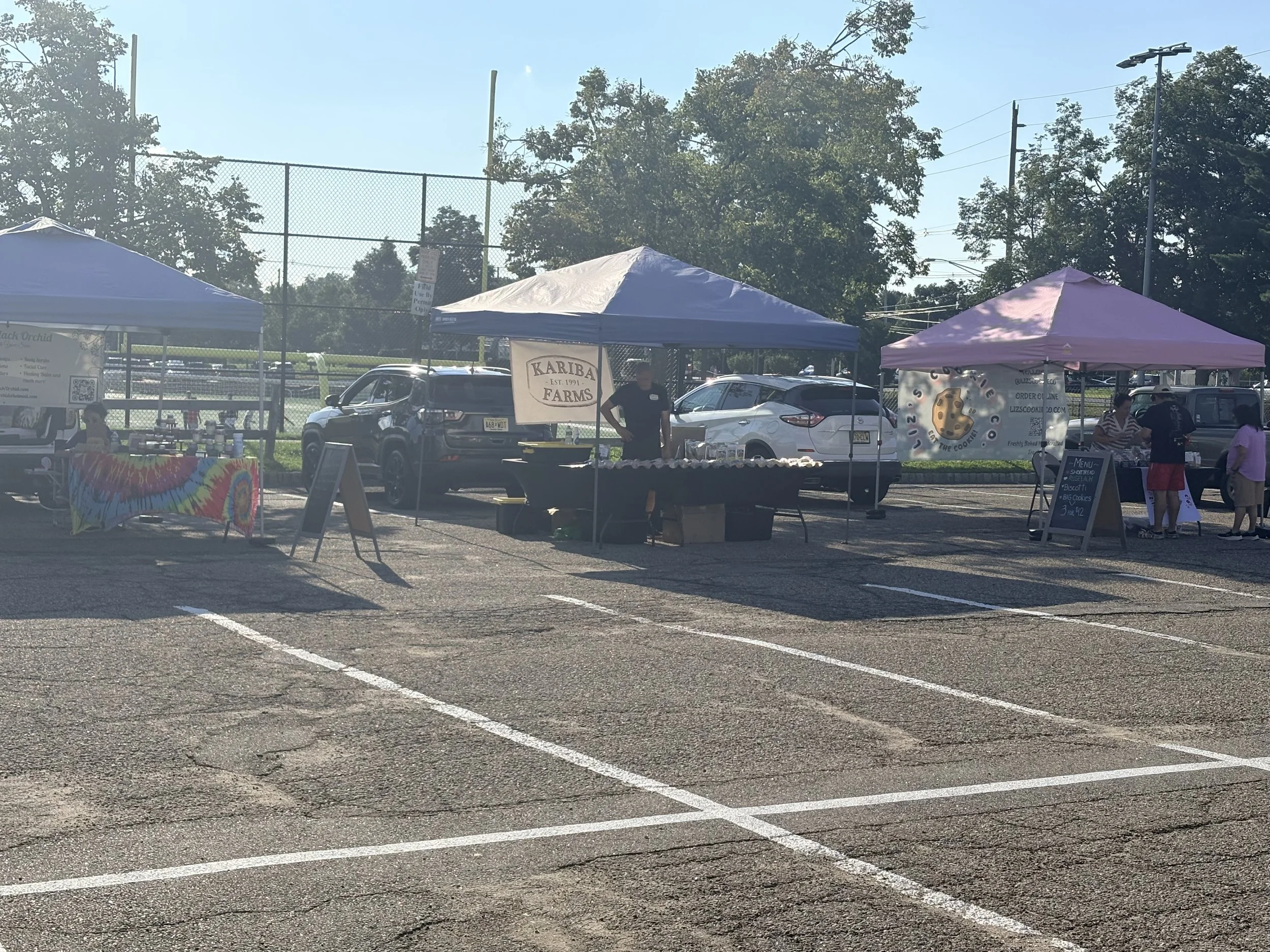 An outdoor market with tents and vendors, parked cars, and people shopping in a parking lot near a sports field with trees and utility poles in the background. Parsippany Farmers market by Hear Here Events 2026 season