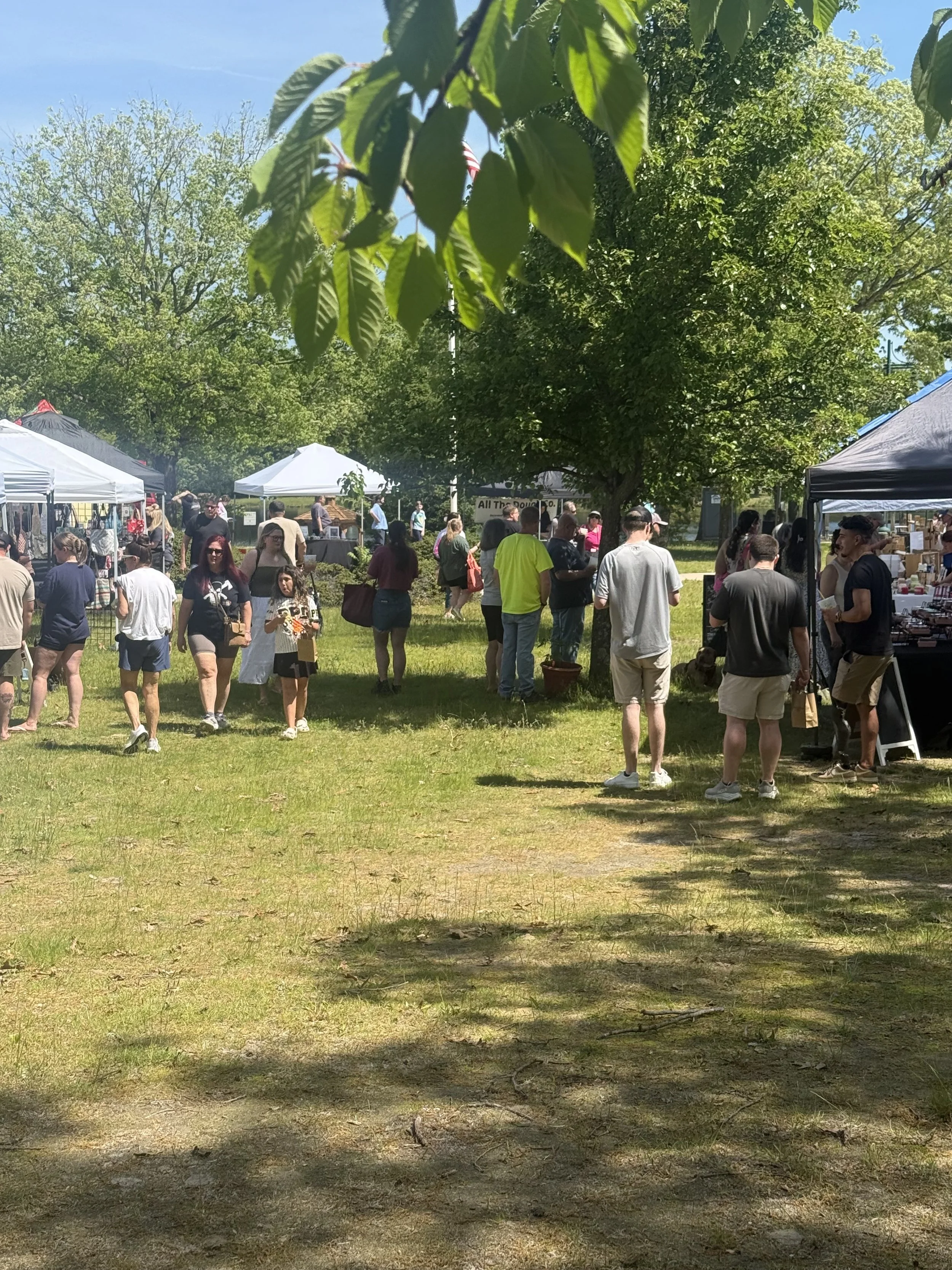 People walking and browsing at an outdoor marketplace with vendor tents, large trees, and a clear, sunny sky. Howell Farmers market by Hear Here