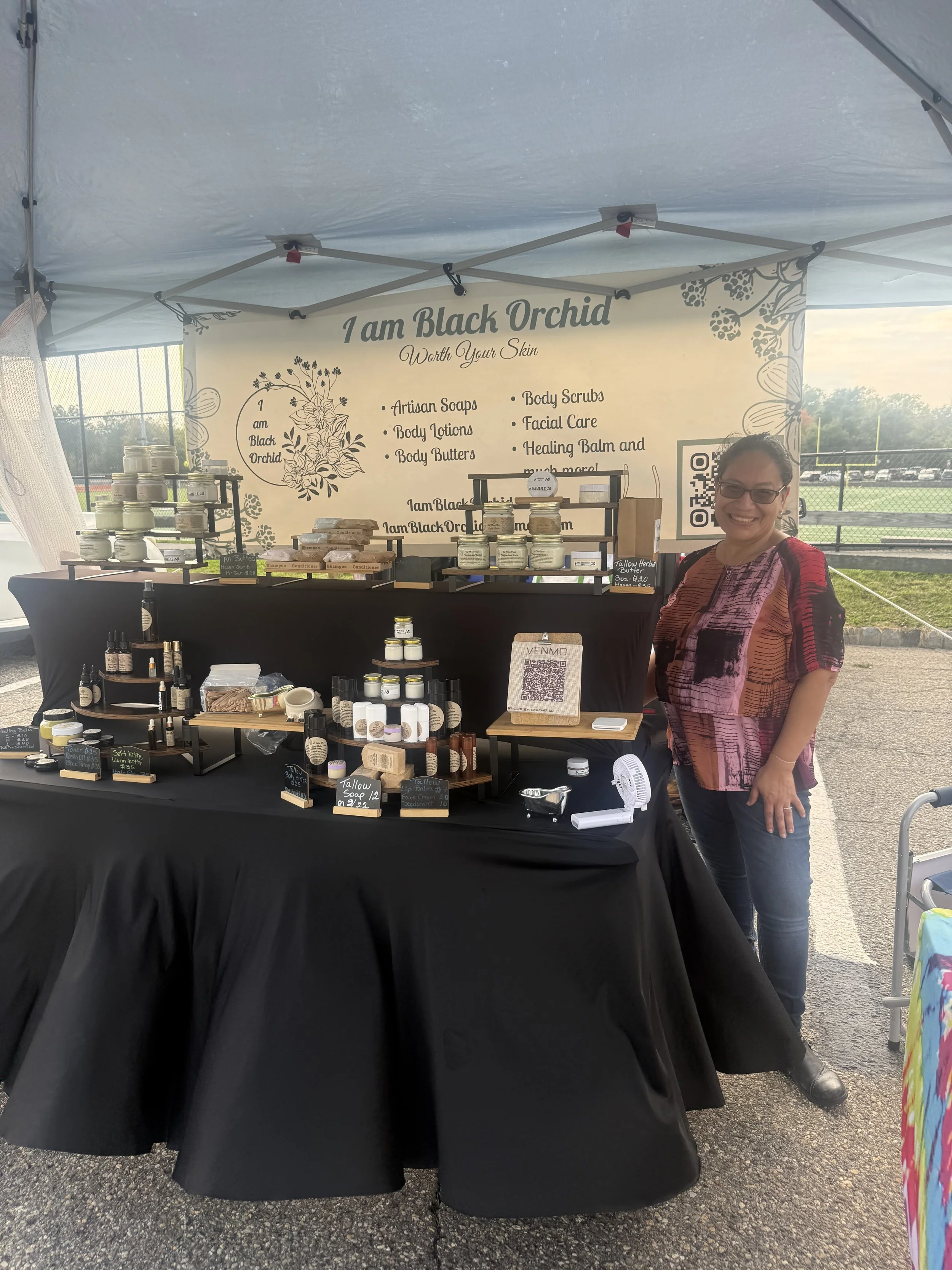 A woman standing next to a booth selling skincare products at an outdoor event. The booth has a sign that reads "I am Black Orchid Worth Your Skin" and displays various jars and bottles of soap, lotion, and other skincare items. Parsippany Farmers 