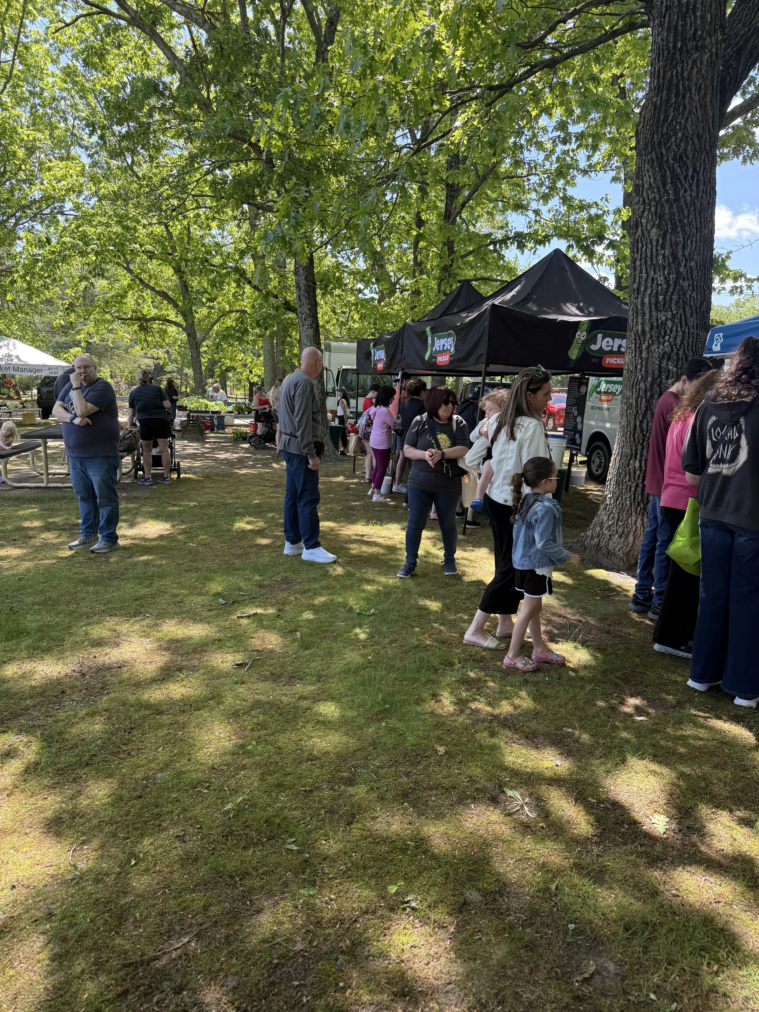 People standing and walking outdoors in a park with large trees and green foliage, some at food stalls or booths. Howell Farmers market by Hear Here Events 2026 season