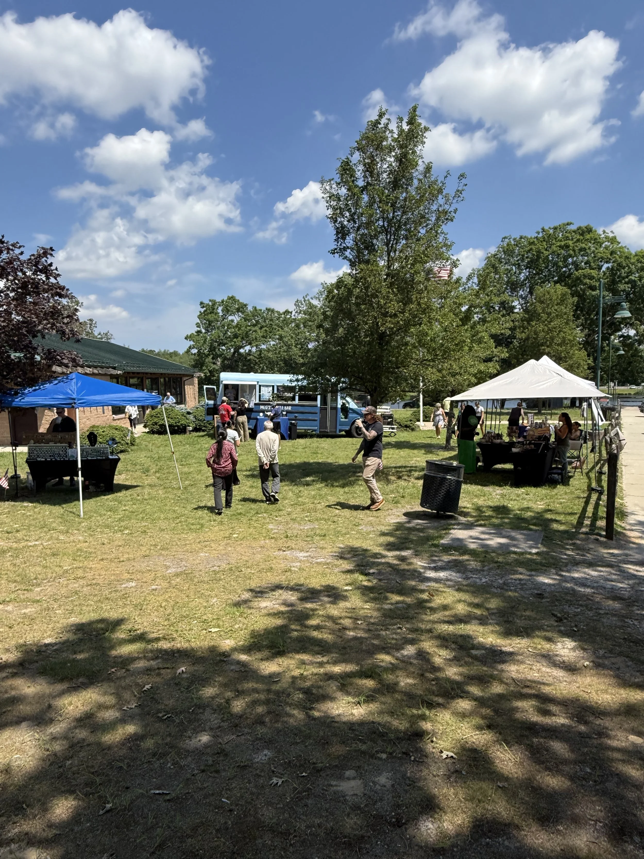 A park scene with people, tents, and food trucks under a blue sky with clouds. There are trees and a grassy area. Howell Farmers market by Hear Here Events 2026 season