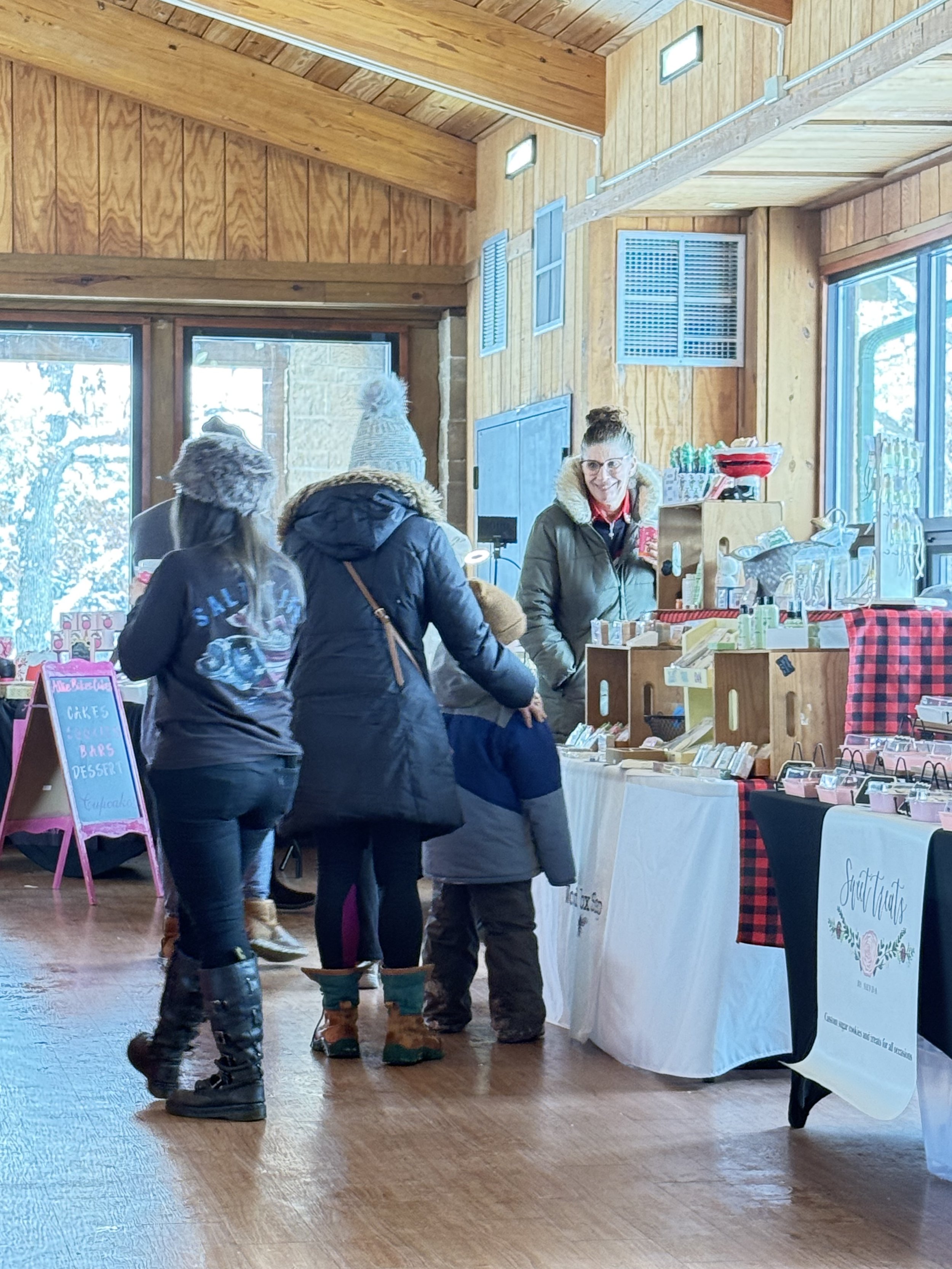 People shopping at a market stall inside a wooden building with high vaulted ceilings, winter clothing, and snow visible outside through large windows. Howell Farmers market by Hear Here Events 2026 season