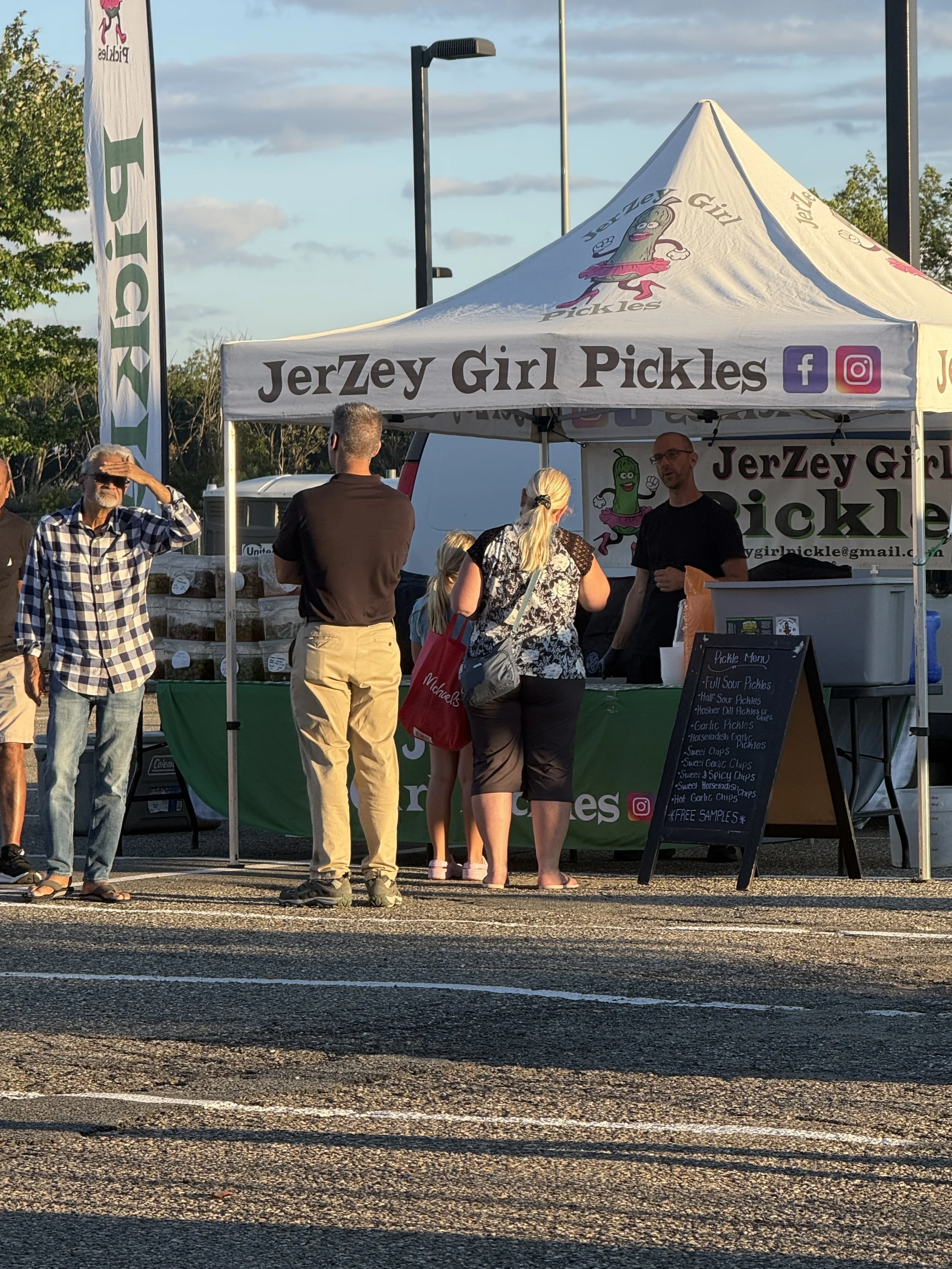 A group of people gathering around a white canopy tent with the sign 'JerZey Girl Pickles' on it. The tent has colorful logos of Facebook and Instagram. There is a chalkboard showing pickle flavors and free samples, with a few trees and a blue sky wi