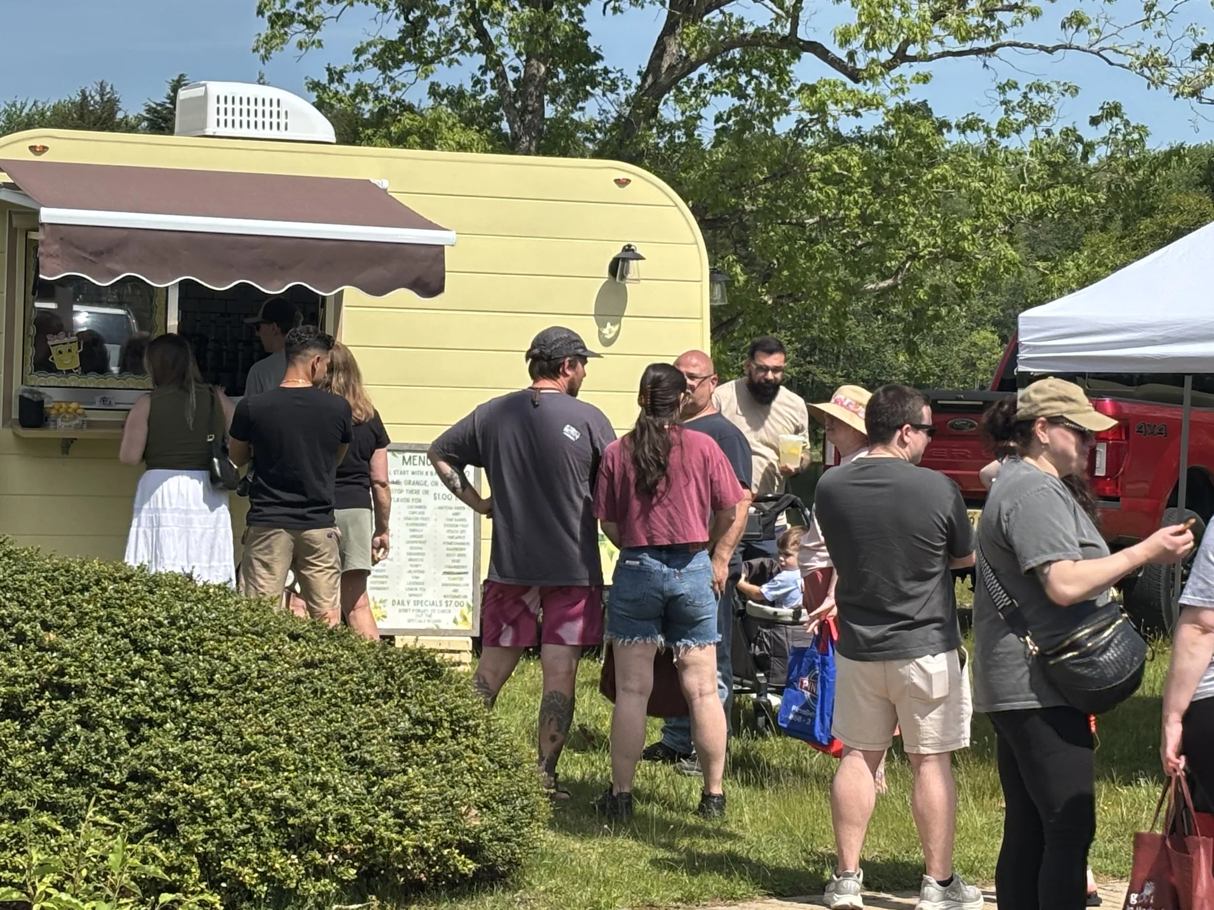 A crowd of people standing in line outdoors next to a yellow food trailer with a brown awning and a menu board. Howell Farmers market by Hear Here
