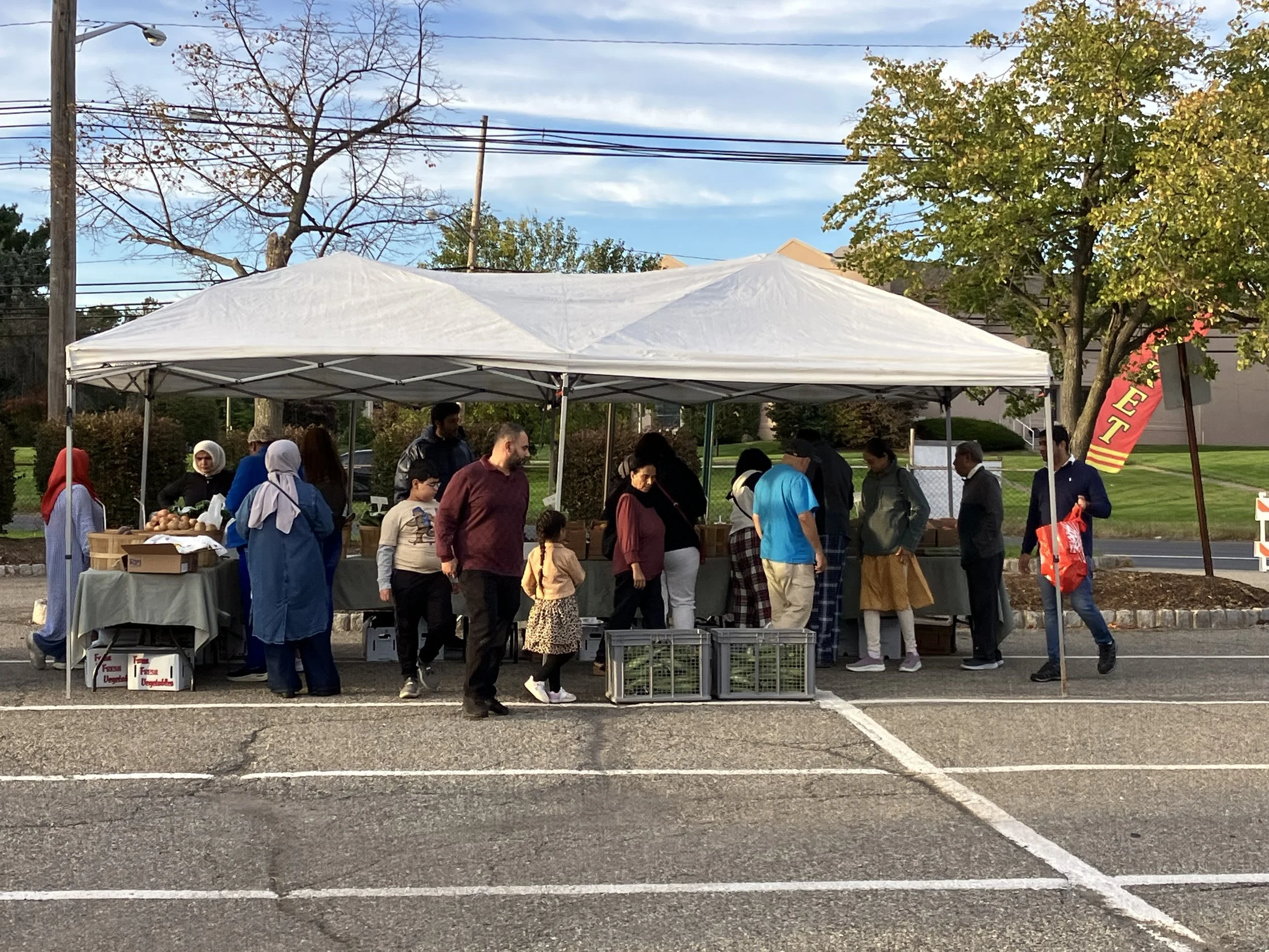 A group of people gathered under a white canopy tent at an outdoor event in a parking lot. There are tables with produce, and a red and yellow banner reading 'SET' is visible on the right side. The background features trees, power lines, and a partly