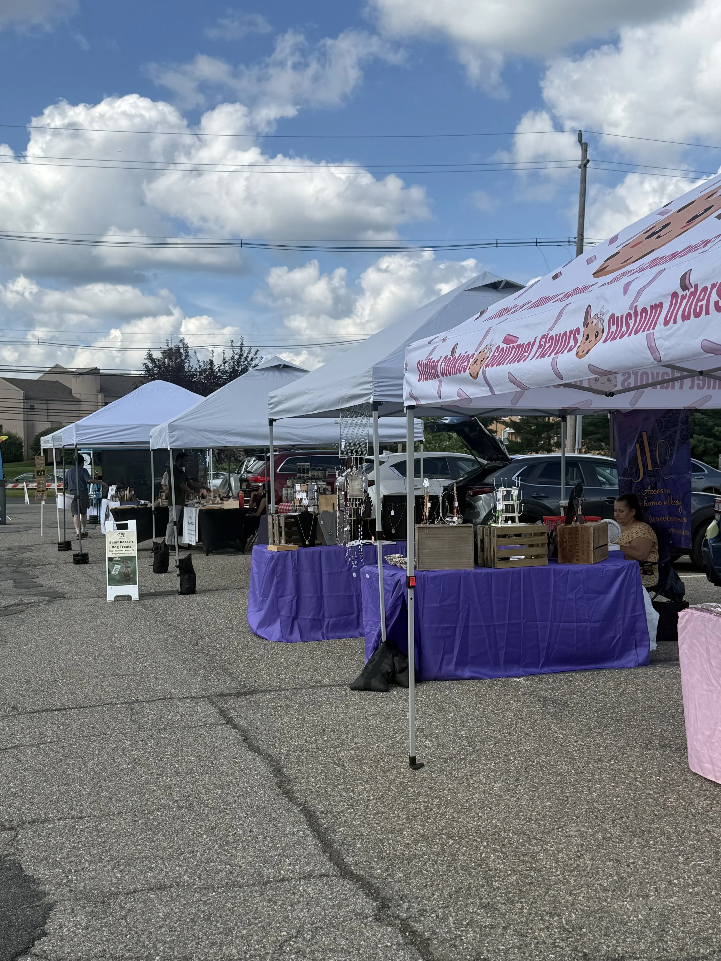 Outdoor market stalls with vendor booths under white tents, displaying jewelry and accessories with a purple tablecloth, in a parking lot on a partly cloudy day. Parsippany Farmers market b y Hear Here Events 2026 season