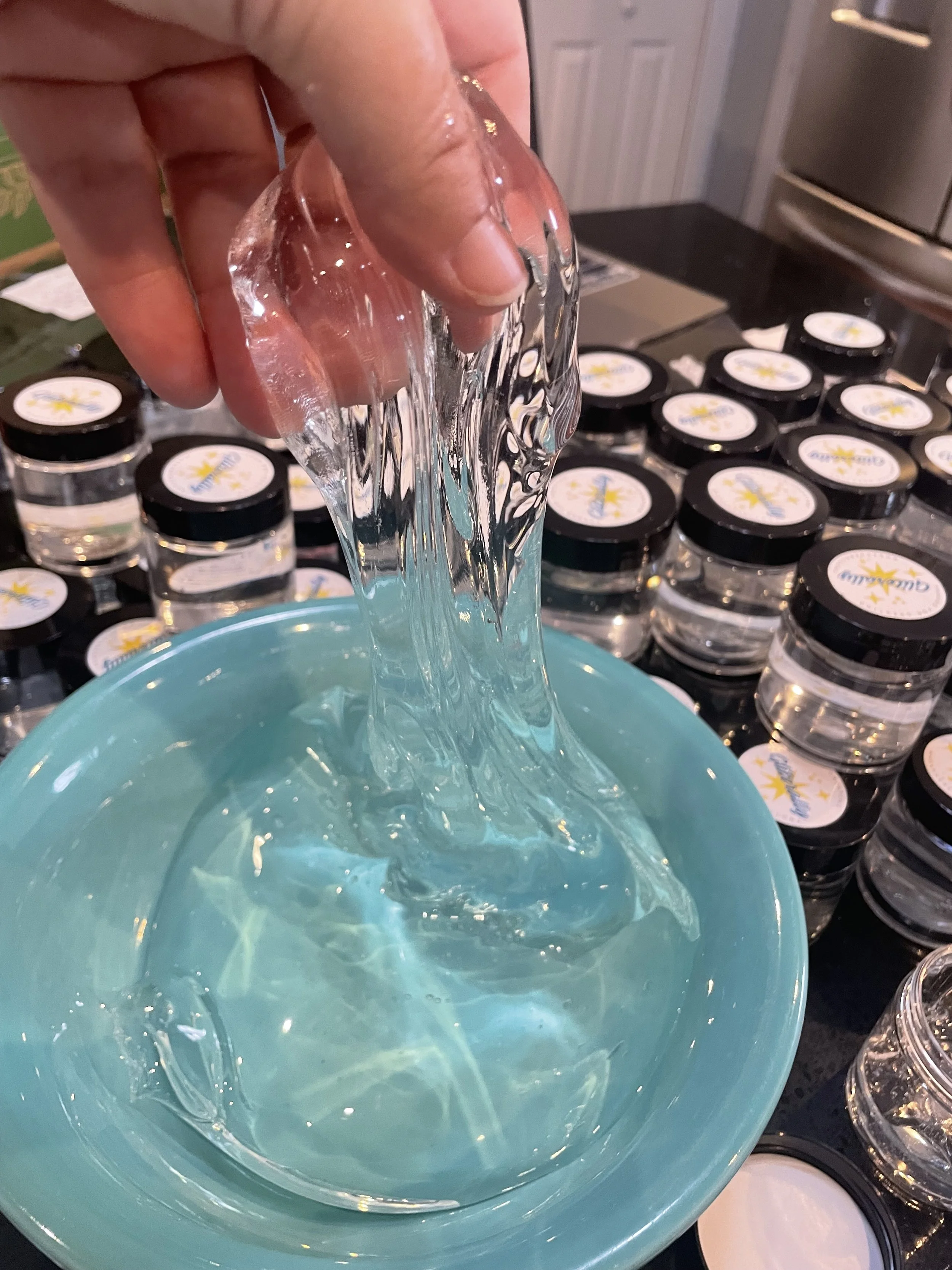 Hand pouring clear, gel-like substance from a small container into a teal bowl, with numerous similar containers in the background.