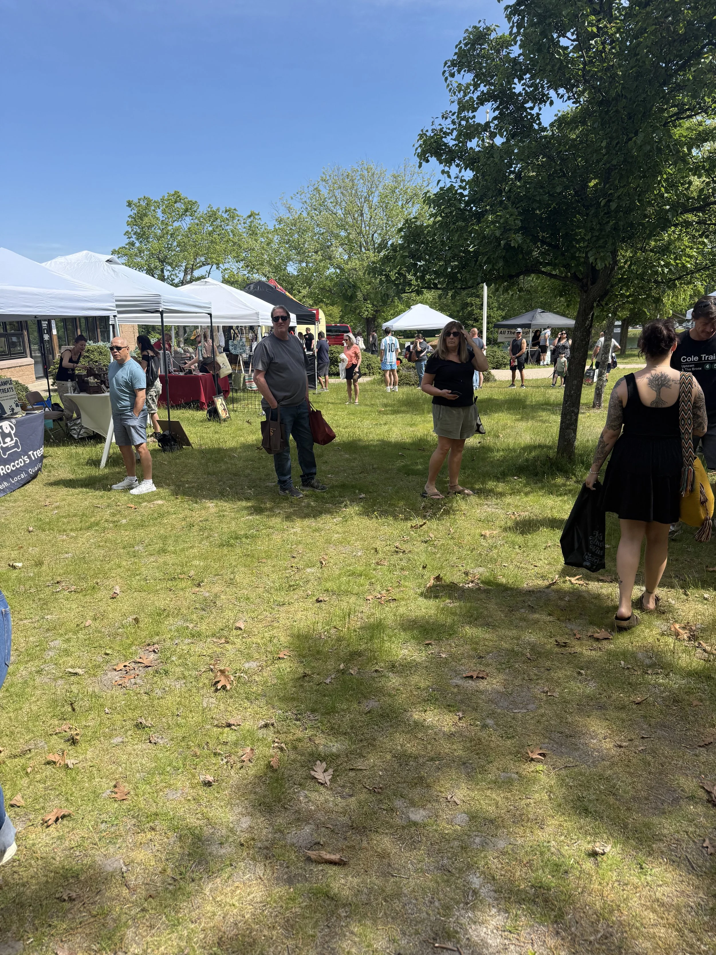 People walking and browsing at outdoor market with white tents under green trees on sunny day. Howell Farmers market by Hear Here