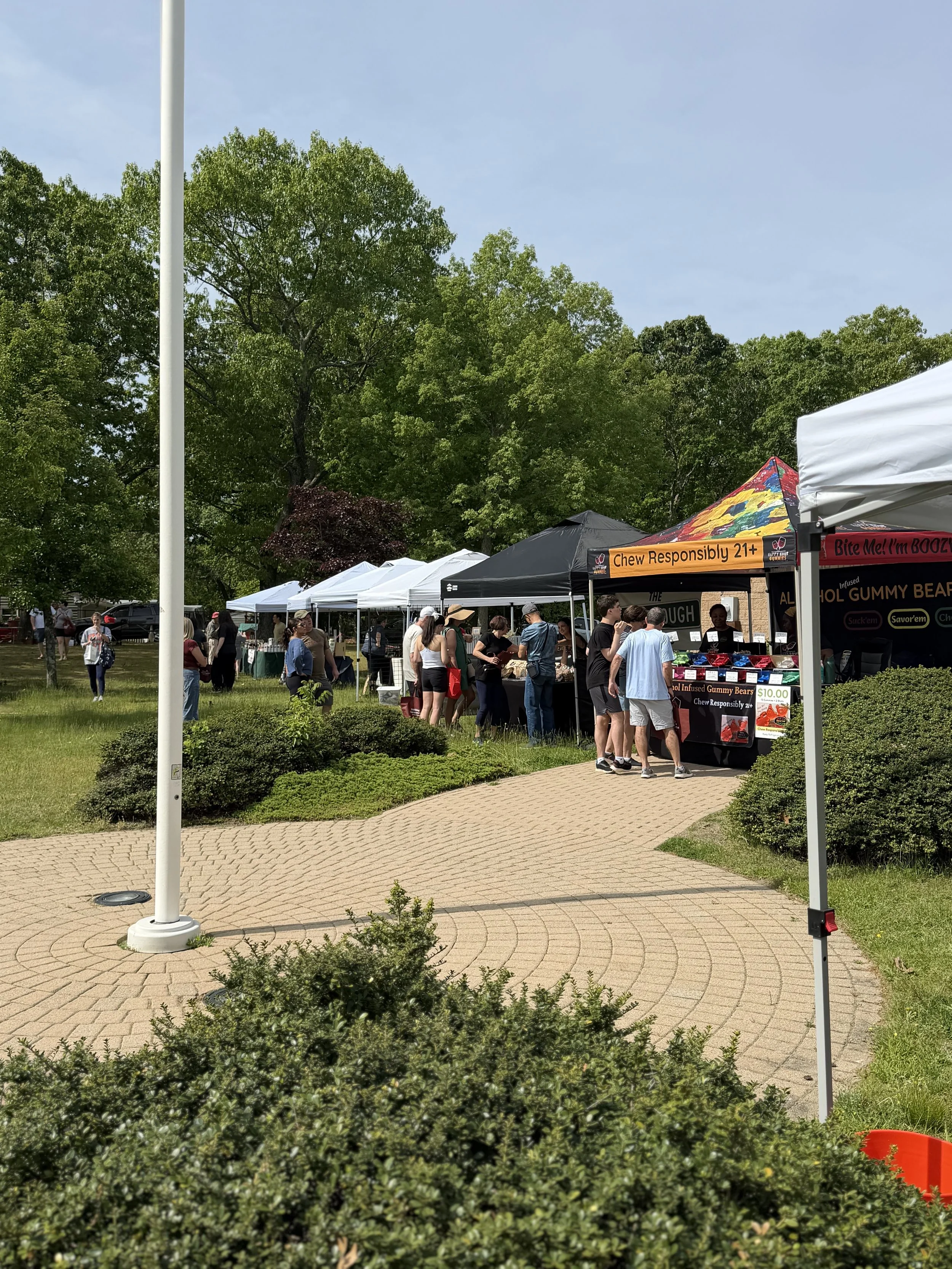 Outdoor market scene with tents, people browsing, and a large banner that reads 'Chew Responsibly 21+'. Howell Farmers market by Hear Here