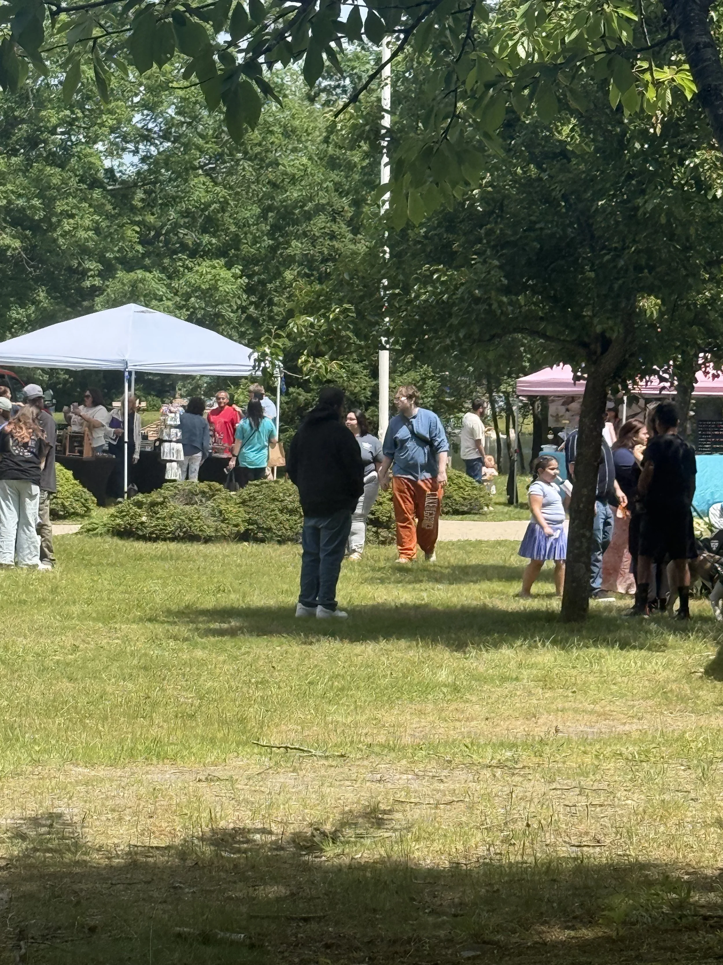 People are gathered outdoors under a large tree during daylight, some are standing, talking, or browsing tables with items, at what appears to be a community event or market. Howell Farmers market by Hear Here