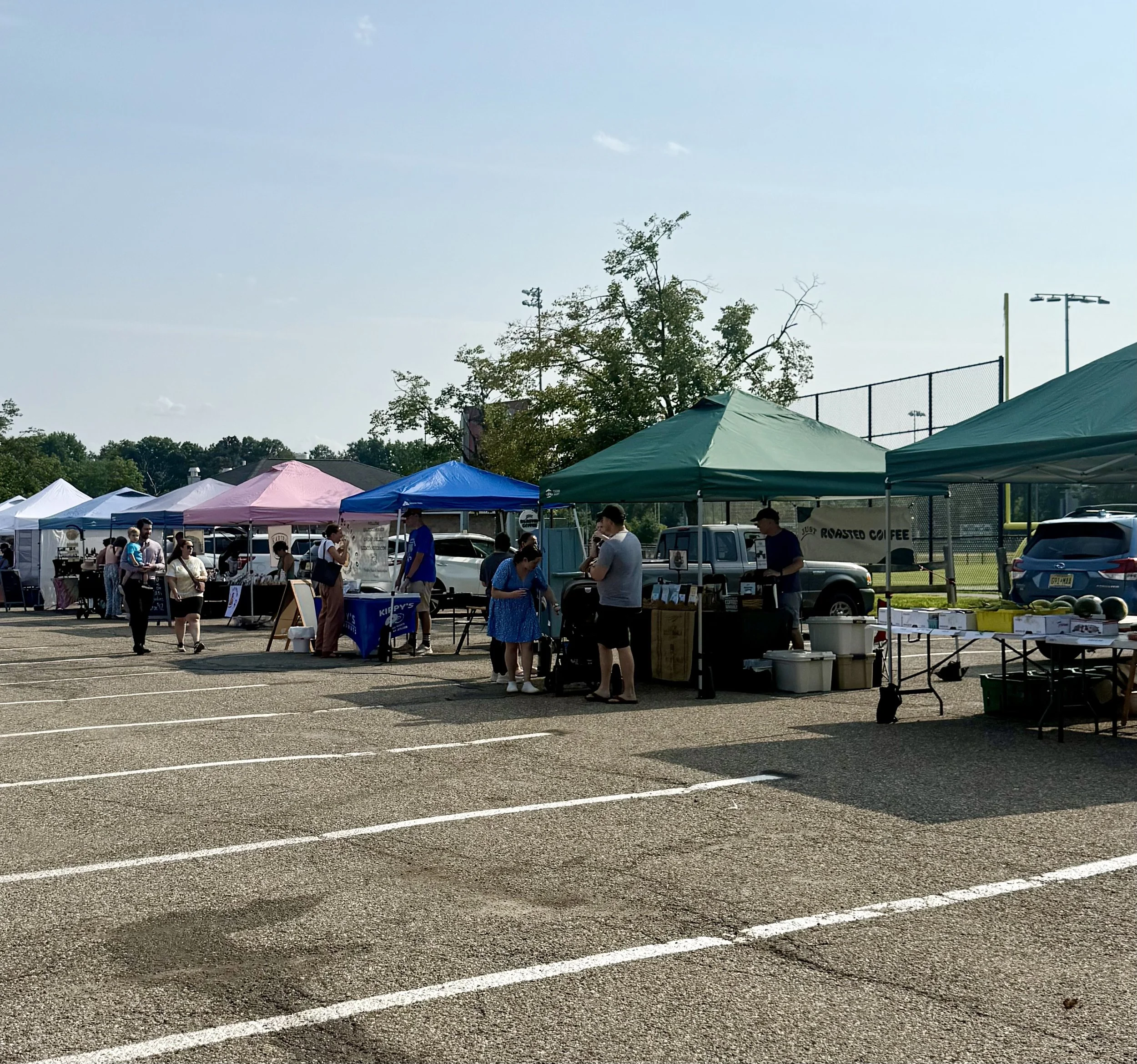 People at an outdoor market or festival with tents, trees, and sports fields visible in the background on a sunny day. Parsippany Farmers market by Hear Here Events 2026 season