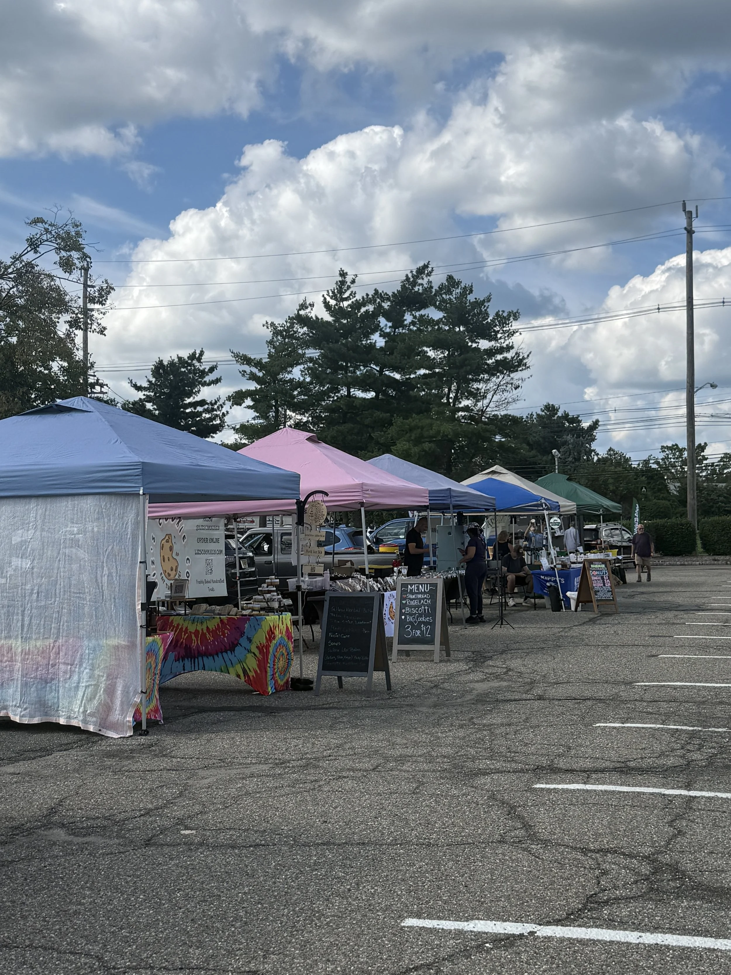 Outdoor market with colorful tents, people browsing and selling goods, parking lot, trees, and cloudy sky in the background. Parsippany Farmers market b y Hear Here Events 2026 season