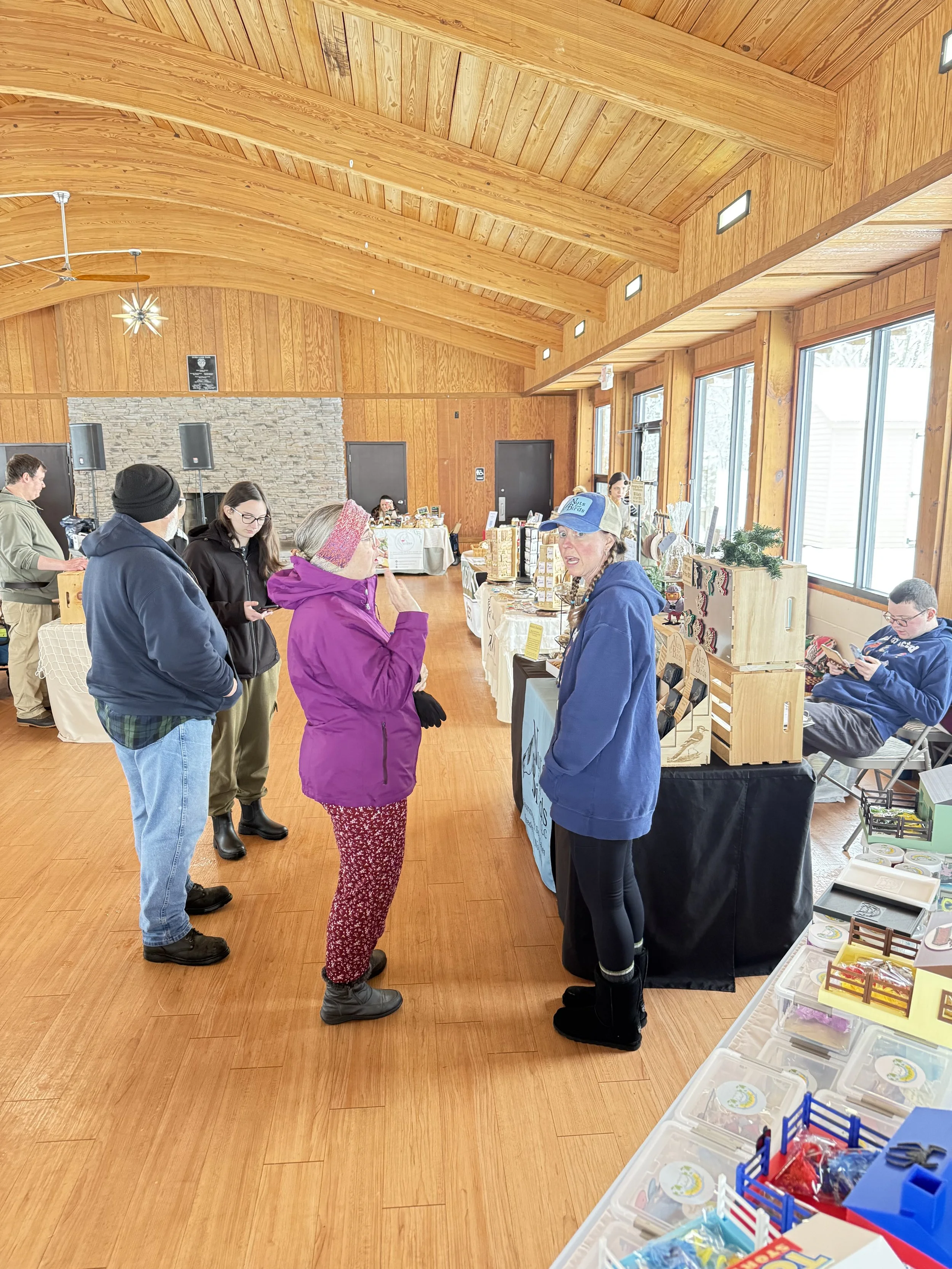People interacting at a craft fair or marketplace inside a wooden building with large windows and a high arched ceiling. Several tables display craft items and products for sale. Howell Farmers market by Hear Here Events 2026 season