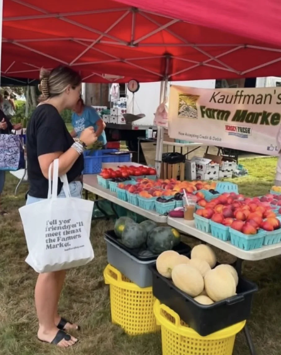 A woman at an outdoor farmer's market stand under a red canopy, looking at fresh peaches and tomatoes for sale. She holds a white tote bag and there are watermelons and honeydew melons on the ground. Allenhurst Farmers market by Hear Here 