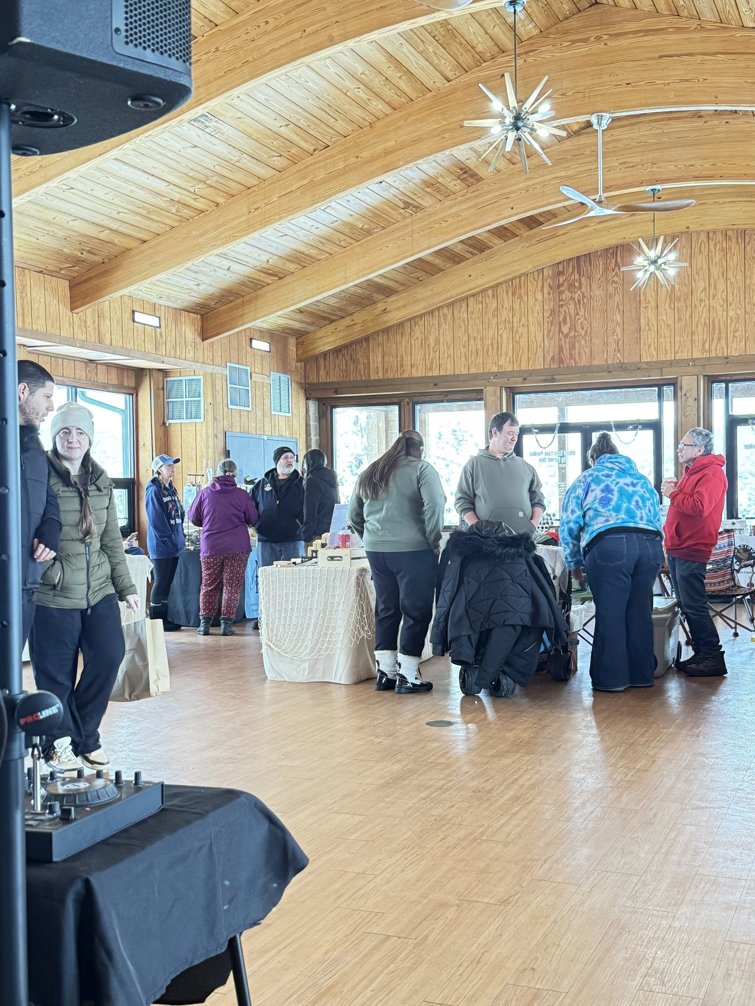 People gathered in a wooden-furnished room with a vaulted ceiling, large windows, and hanging star-shaped lights, at what appears to be a market or fair. Howell Farmers market by Hear Here Events 2026 season