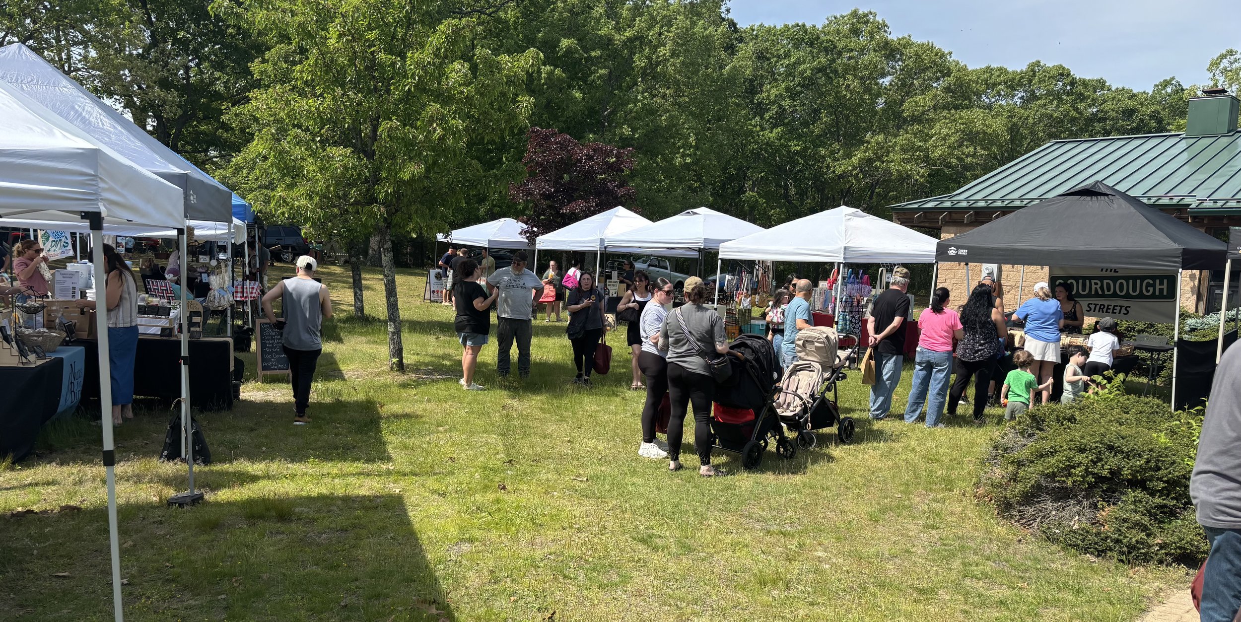 People browsing outdoor vendors at a market with white tents on a sunny day, surrounded by trees. Howell Farmers market by Hear Here