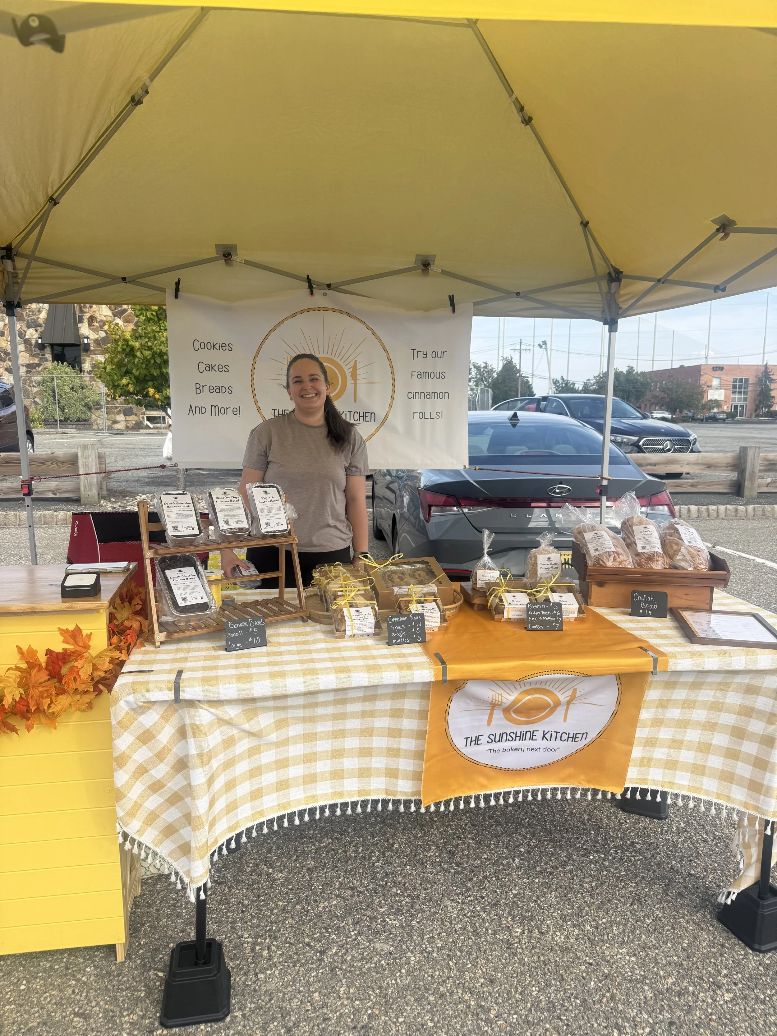 A woman standing behind a table at an outdoor market stall with baked goods like cinnamon rolls, banana bread, and cookies, Parsippany Farmers market by Hear Here Events 2026 season