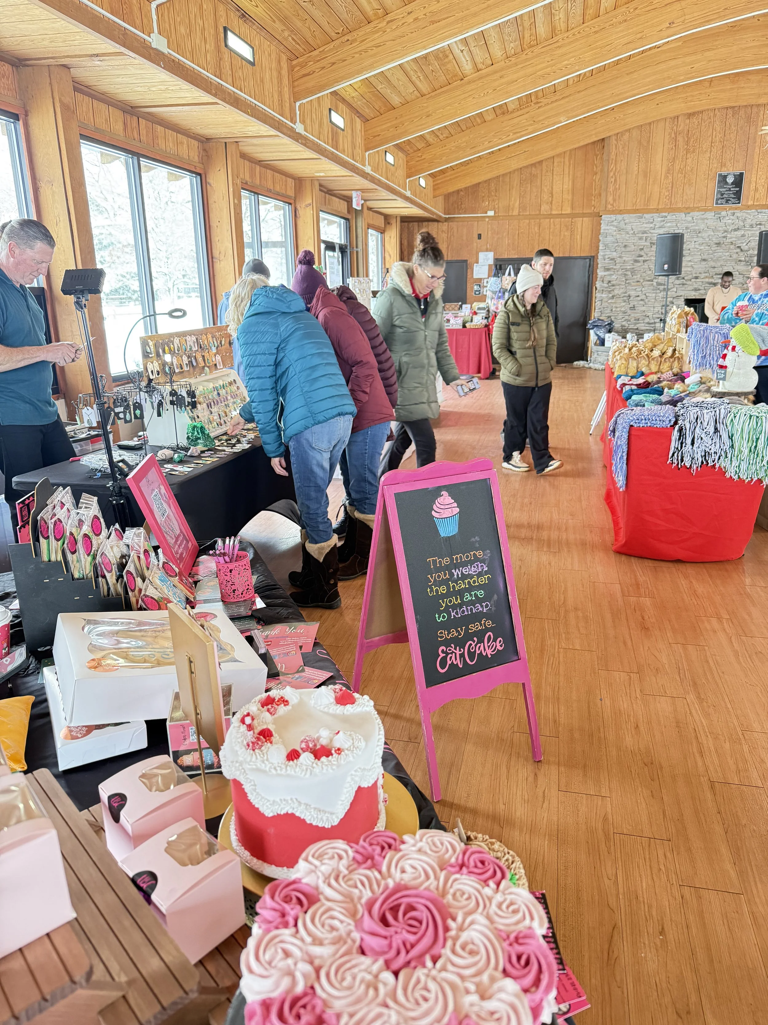 Indoor market or craft fair with various tables displaying jewelry, baked goods, and holiday-themed items, under wooden walls and ceiling with large windows. Howell Farmers market by Hear Here Events 2026 season