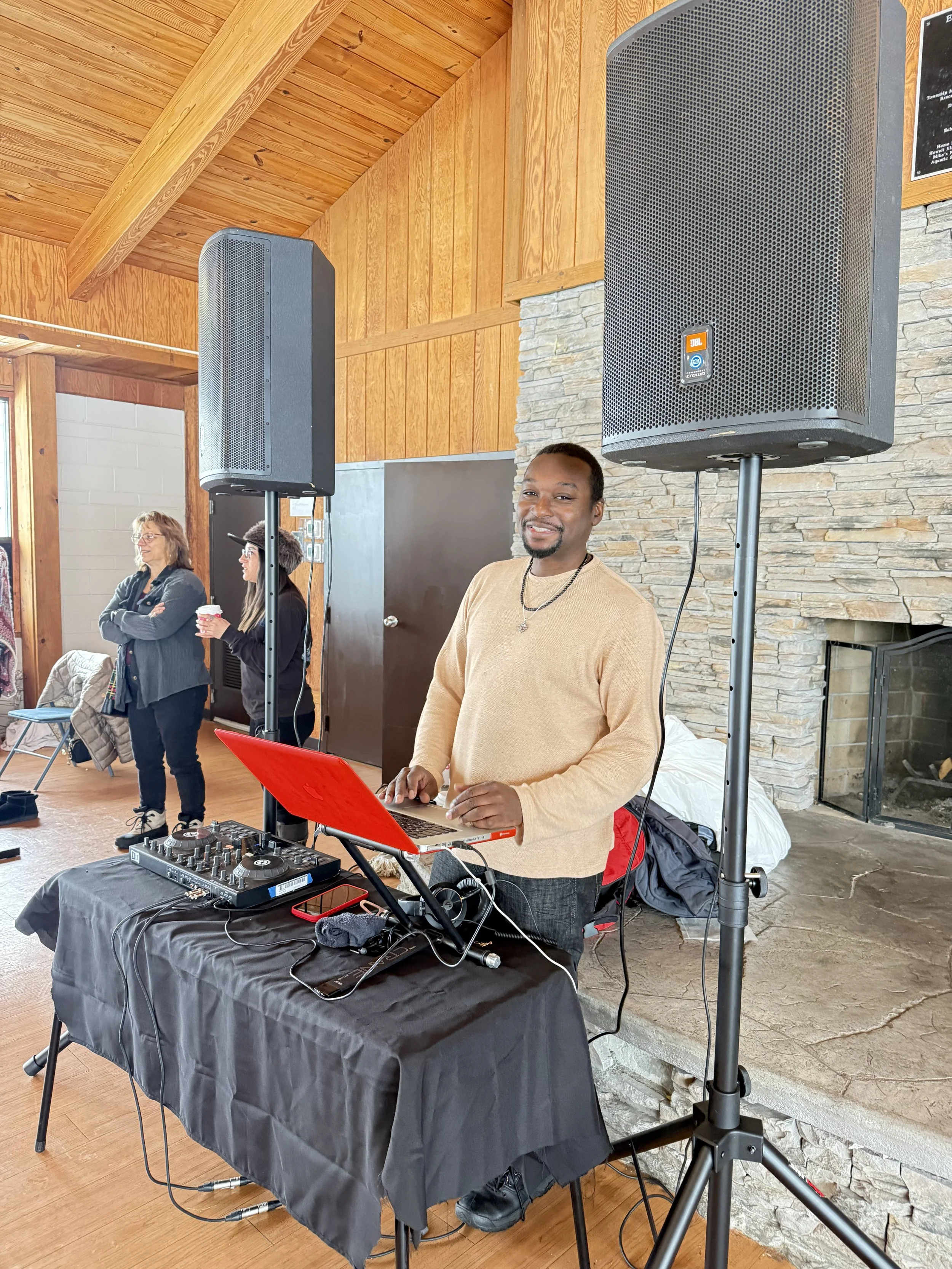 A smiling man standing behind DJ equipment, including a mixer and laptop, with large speakers on stands around him, at an indoor gathering with wooden walls and people in the background. Howell Farmers market by Hear Here Events 2026 season