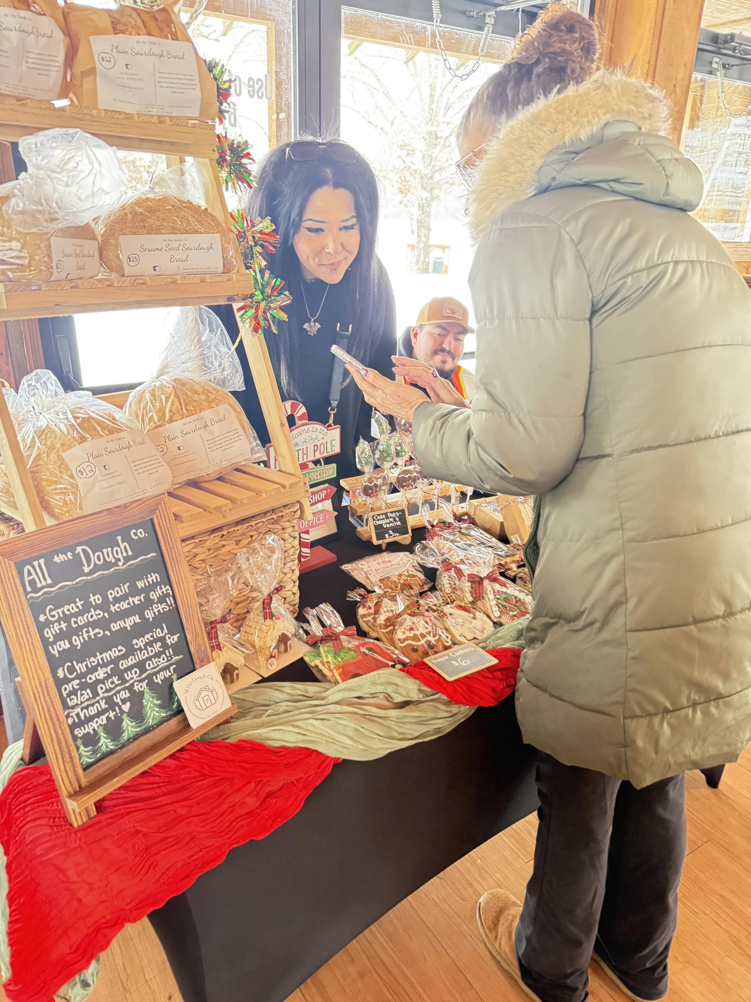 A man in a gray jacket and boots is purchasing baked goods from a woman at a holiday market stall. The stall displays various bread, cookies, and festive decorations, with a sign promoting gift cards Howell Farmers market by Hear Here Events 2026 