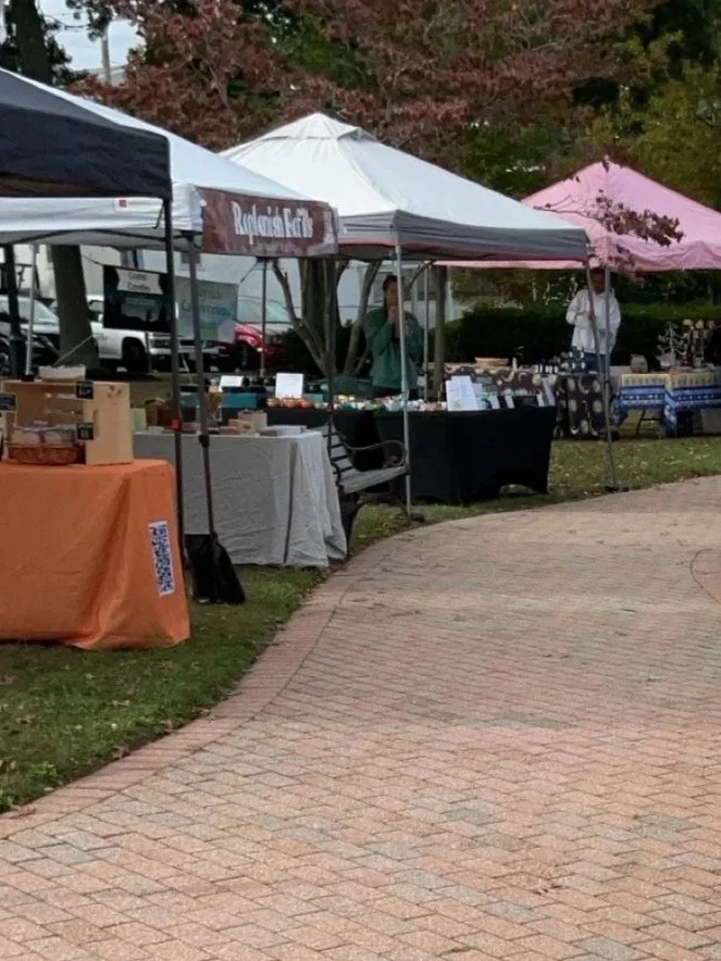 Outdoor farmers market with vendor tents and tables displaying various goods, under trees with red and green leaves.