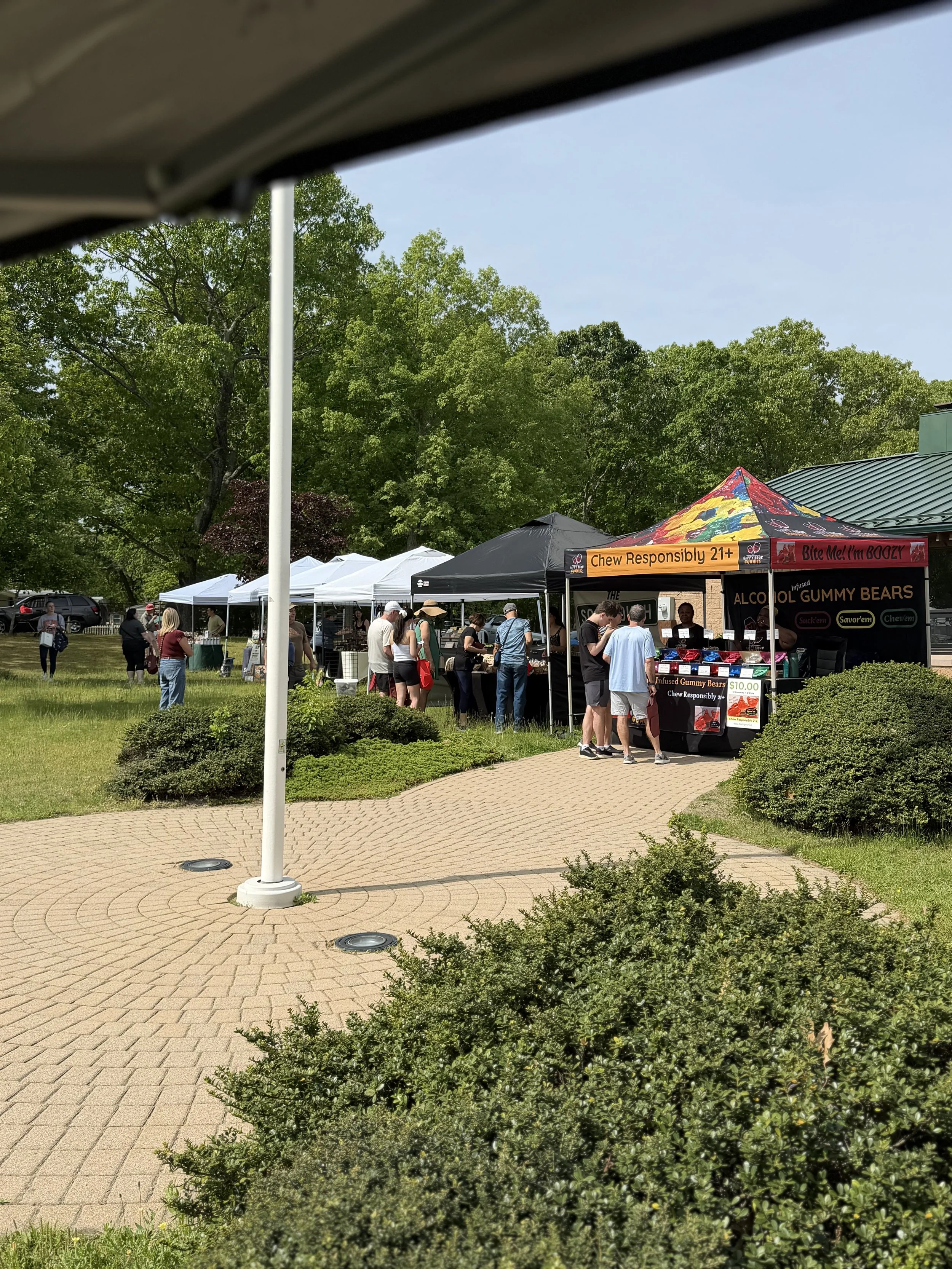 Outdoor market scene with tents and people, including a black tent with an orange sign that says 'Chew Responsibly 21+' and a pink sign with 'Bite Me! I'm Boozy', surrounded by trees and landscaping. Howell Farmers market by Hear Here Events 2026 