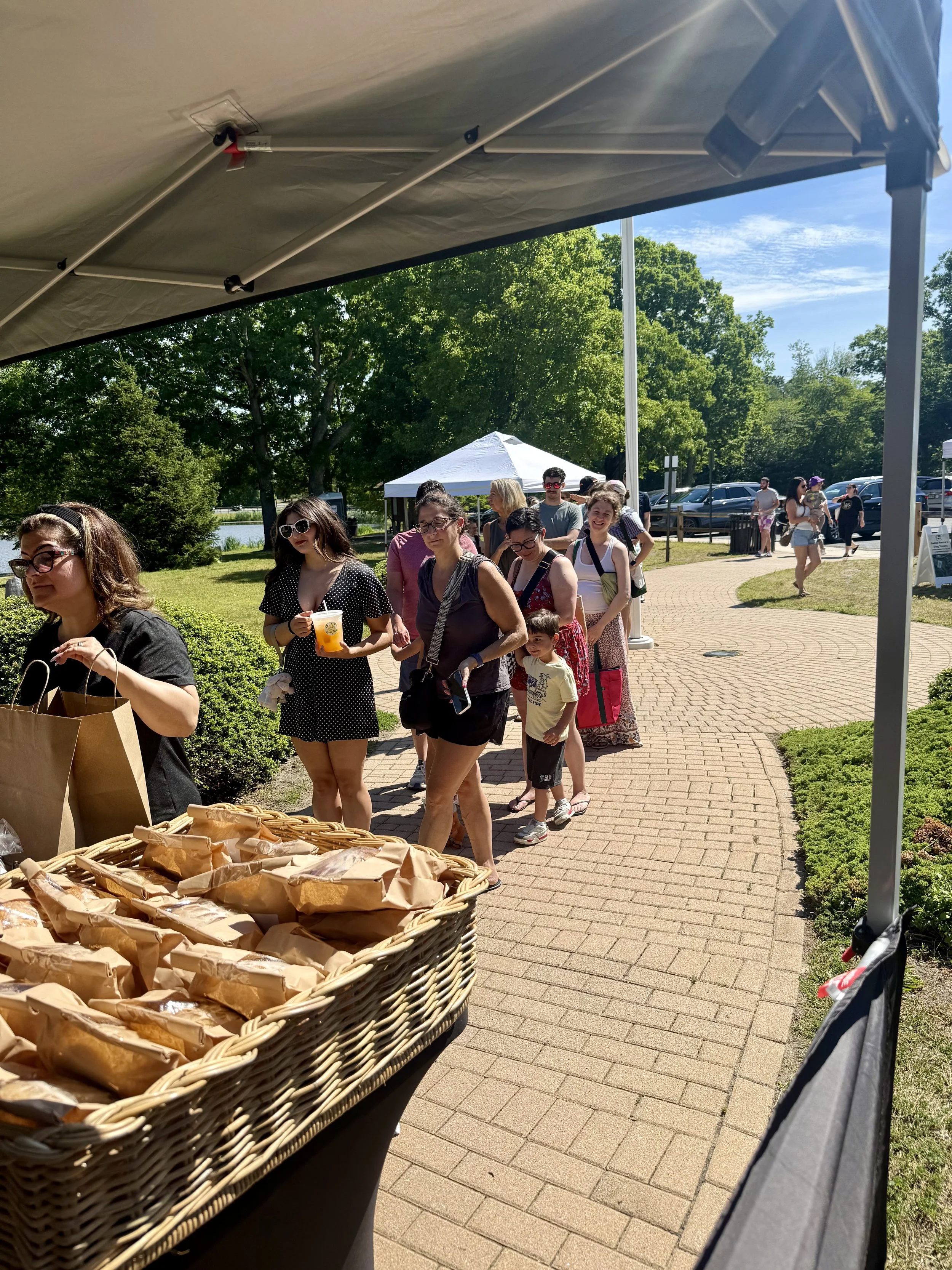 People standing in line outdoors under a shade canopy, with a basket of bread in the foreground, in a park-like setting with trees, a white tent, and a parking lot in the background. Howell Farmers market by Hear Here Events 2026 season
