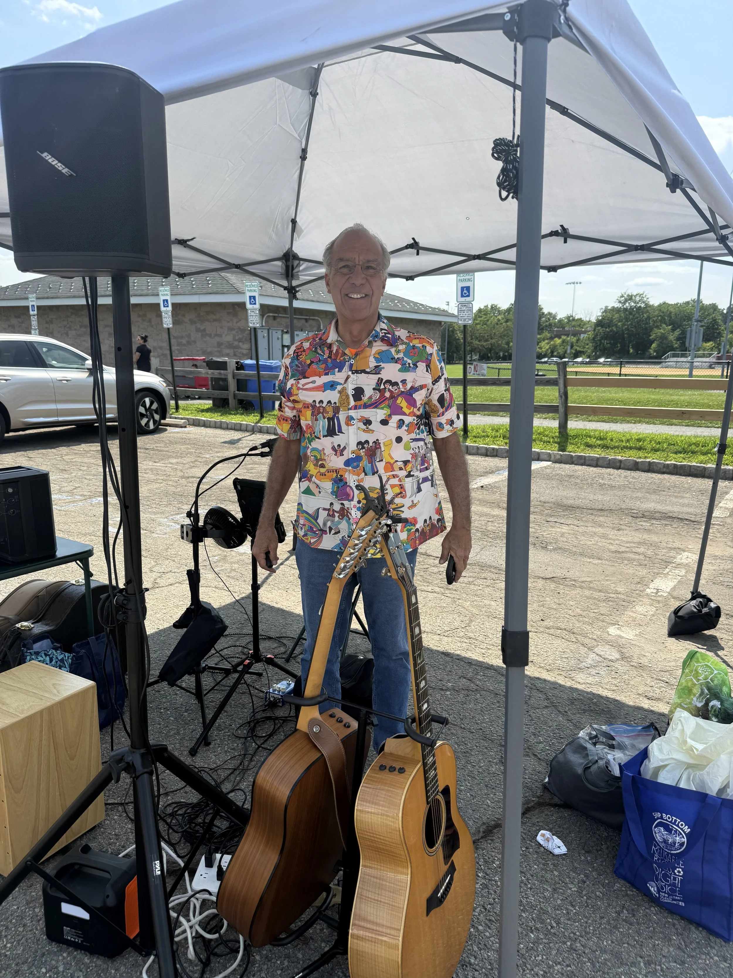 An older man standing under a white canopy tent outdoors, smiling, wearing a colorful short-sleeved shirt with a dinosaur pattern. He is holding a smartphone in one hand, with two acoustic guitars in front of him. There are audio equipment, cables, a
