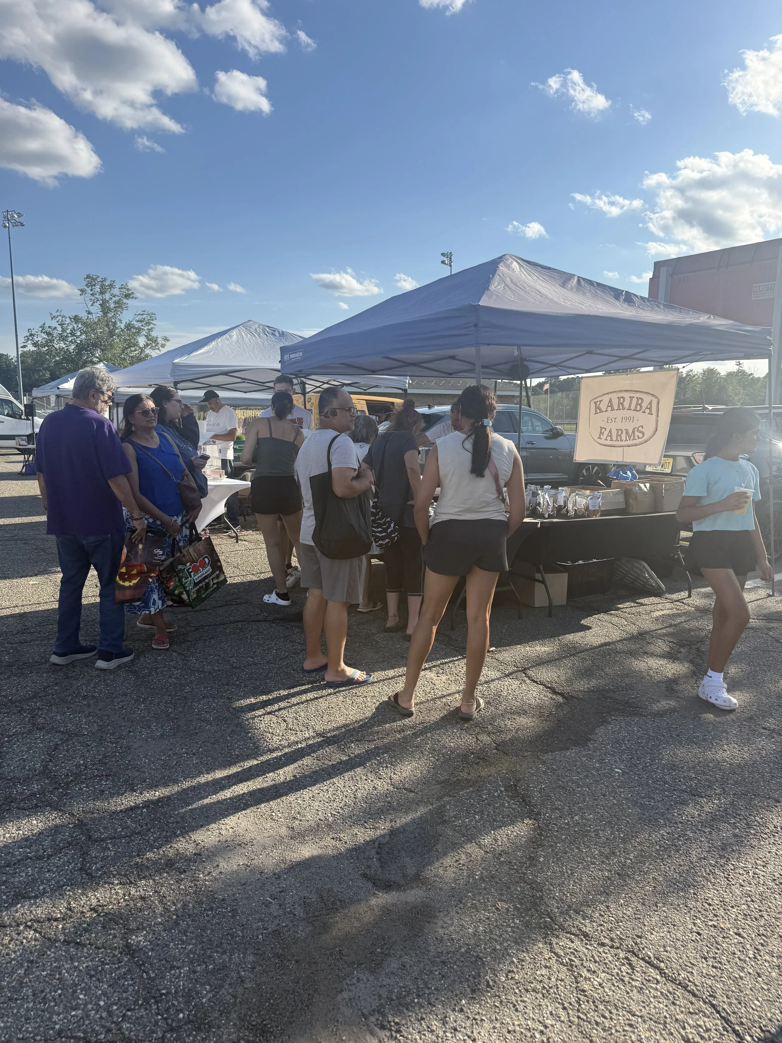 People shopping at an outdoor market stall under a white canopy with a sign that reads 'Kariba Farms,' on a sunny day with blue sky and scattered clouds.Parsippany Farmers market b y Hear Here Events 2026 season