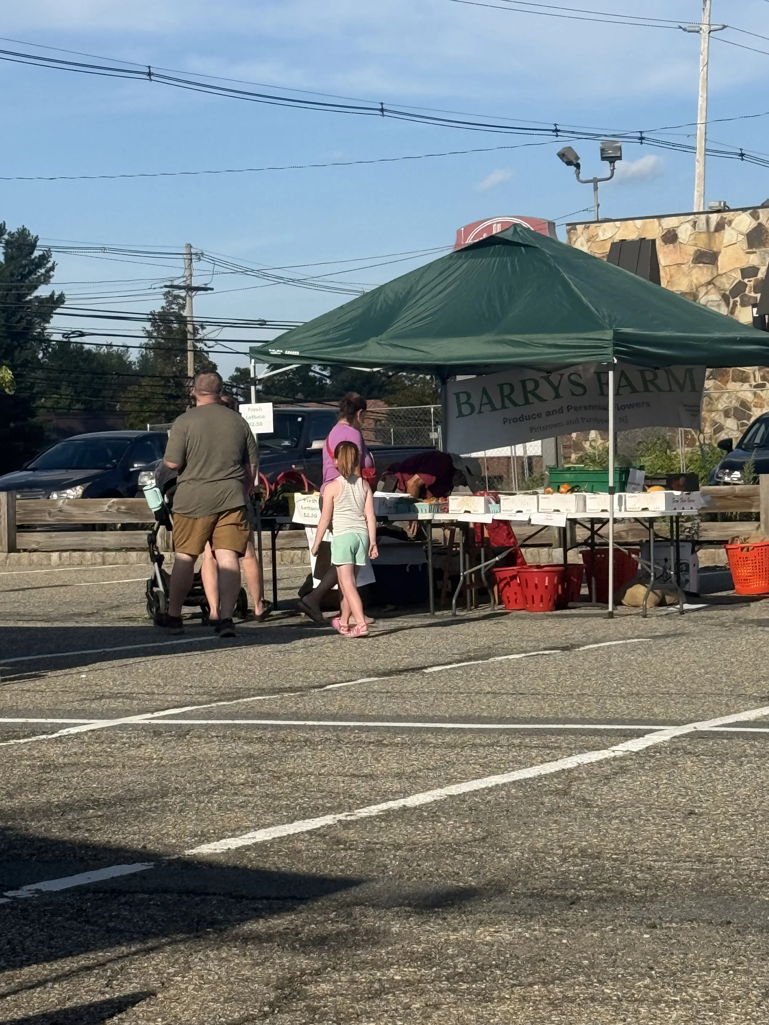 People shopping at an outdoor farmers market with a green tent that has a sign for Barry’s Farm, produce, and flowers, in a parking lot with cars and power lines in the background. Parsippany Farmers market by Hear Here Events 2026 season