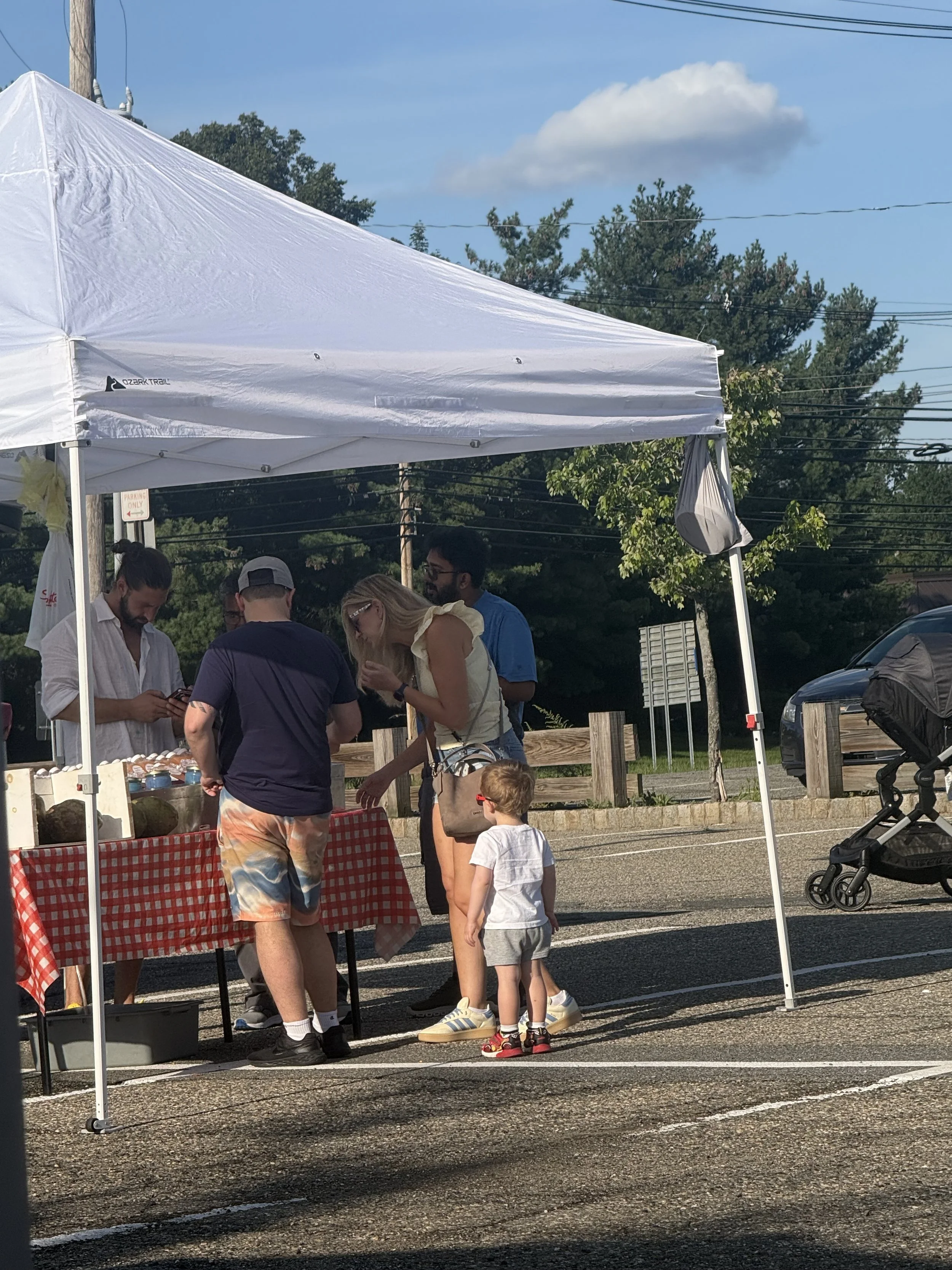 People gathered at a booth with a red and white checkered tablecloth under a white canopy at an outdoor event on a sunny day, with trees and power lines in the background, and a stroller nearby. Parsippany Farmers market by Hear Here Events 2026