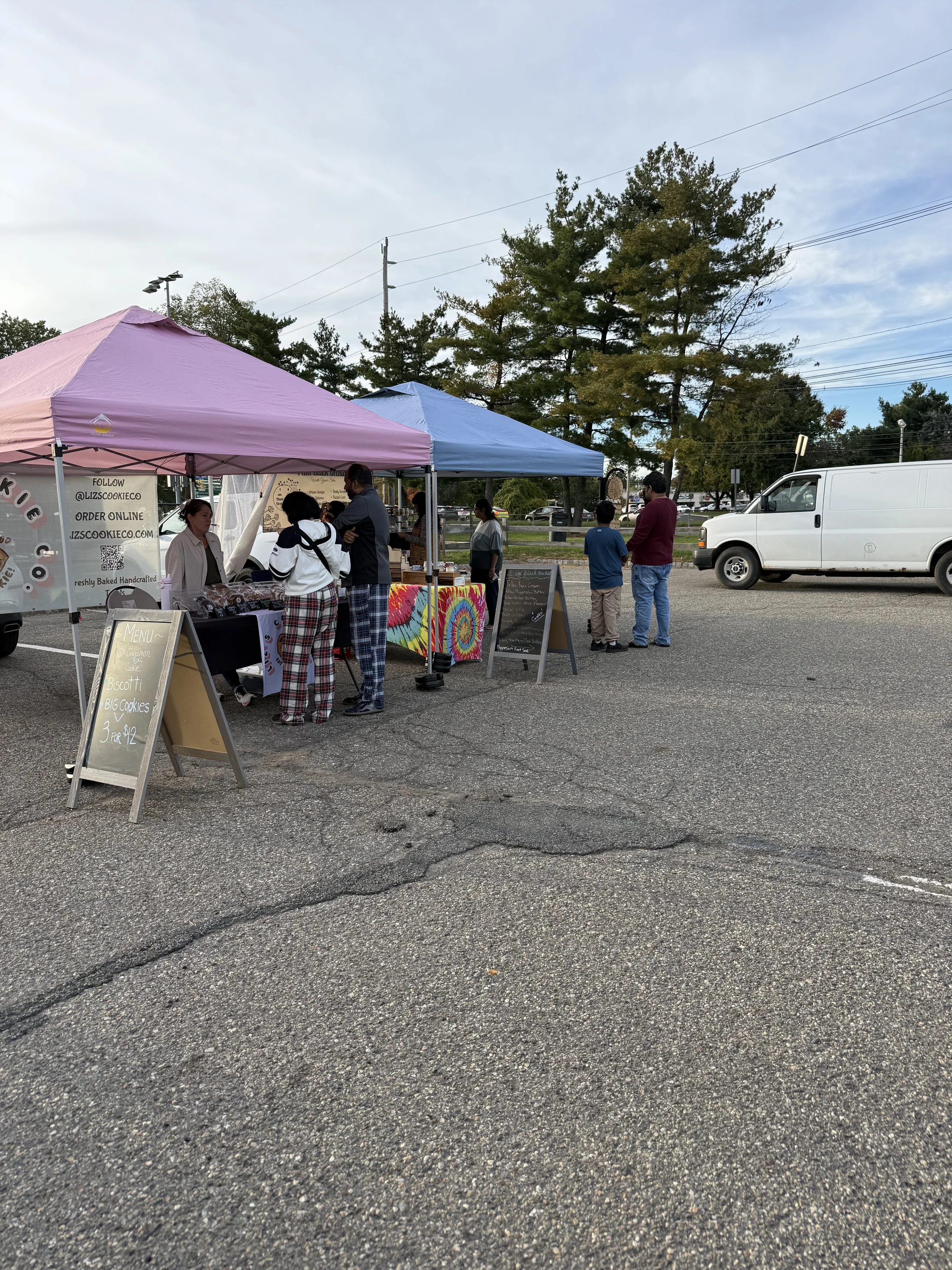 Outdoor market scene with two canopies, people shopping, and a white van parked nearby on a cracked asphalt lot, with trees and power lines in the background. Parsippany Farmers market by Hear Here Events 2026 season