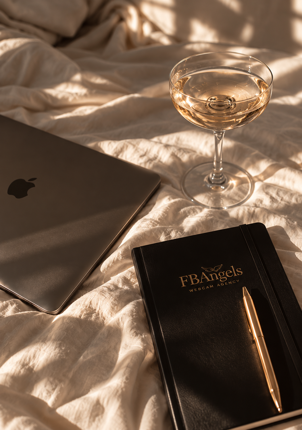 A champagne glass, laptop and black FBAngels notebook with a gold pen arranged on cream bedding in warm natural sunlight.