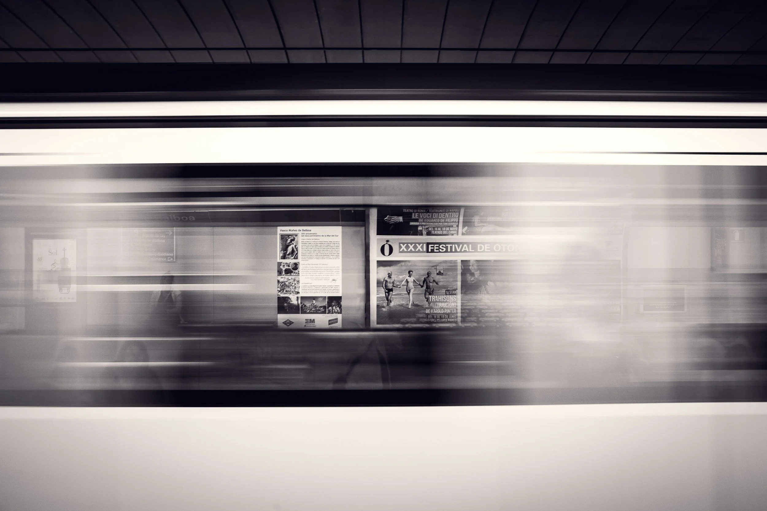 A blurred image of a subway or train station platform with a train passing by quickly. There are posters on the wall in the background, including one advertising a festival, and the overall scene is in black and white.