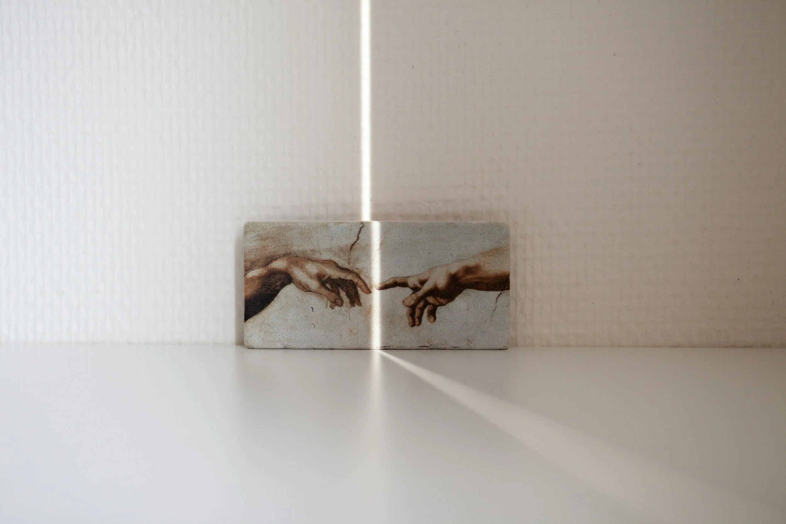 A small rectangular object with an image of Michelangelo's 'The Creation of Adam' on it, positioned against a white wall with light creating a shadow.