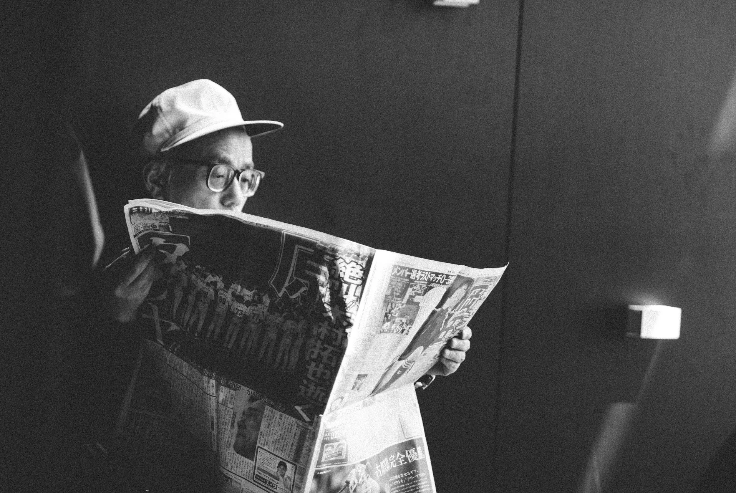 A person wearing glasses and a cap, sitting and reading a newspaper in a dimly lit room.